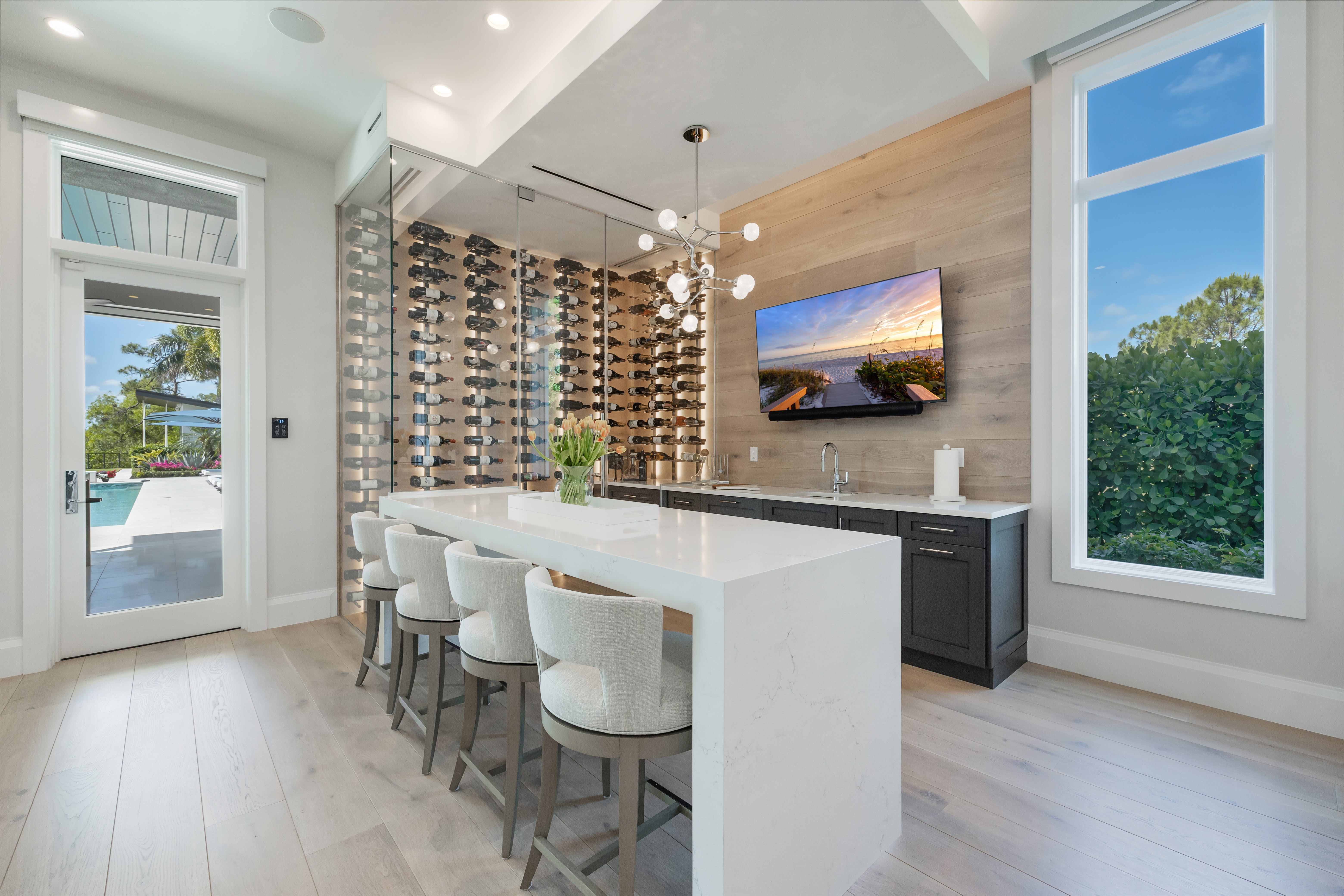 Modern kitchen with a long white island, four upholstered stools, glass enclosed wine rack, wall-mounted TV, and large windows showing greenery and pool outside.