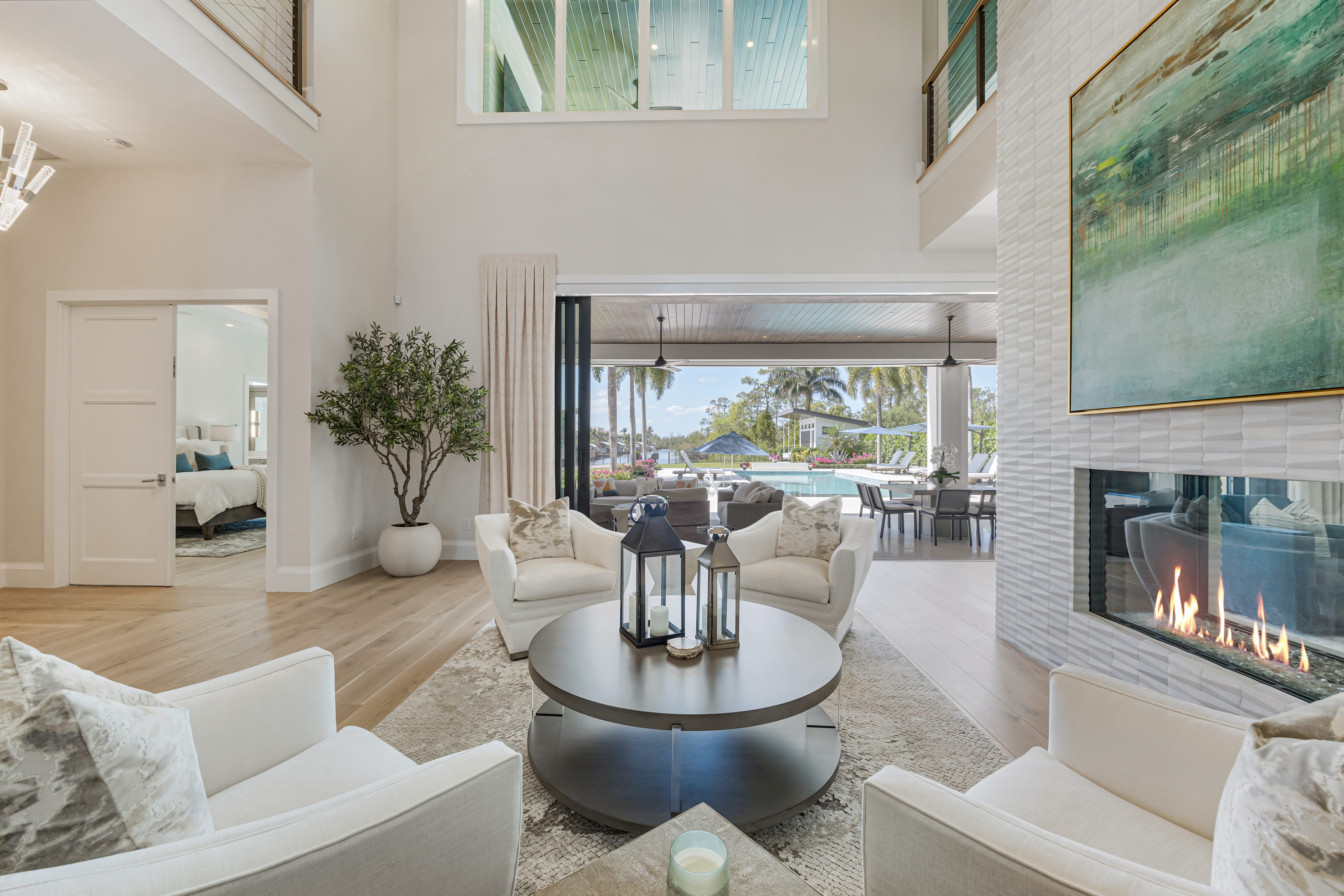 Modern living room with white armchairs, round coffee table with lanterns, fireplace, and open view to a pool and patio area.