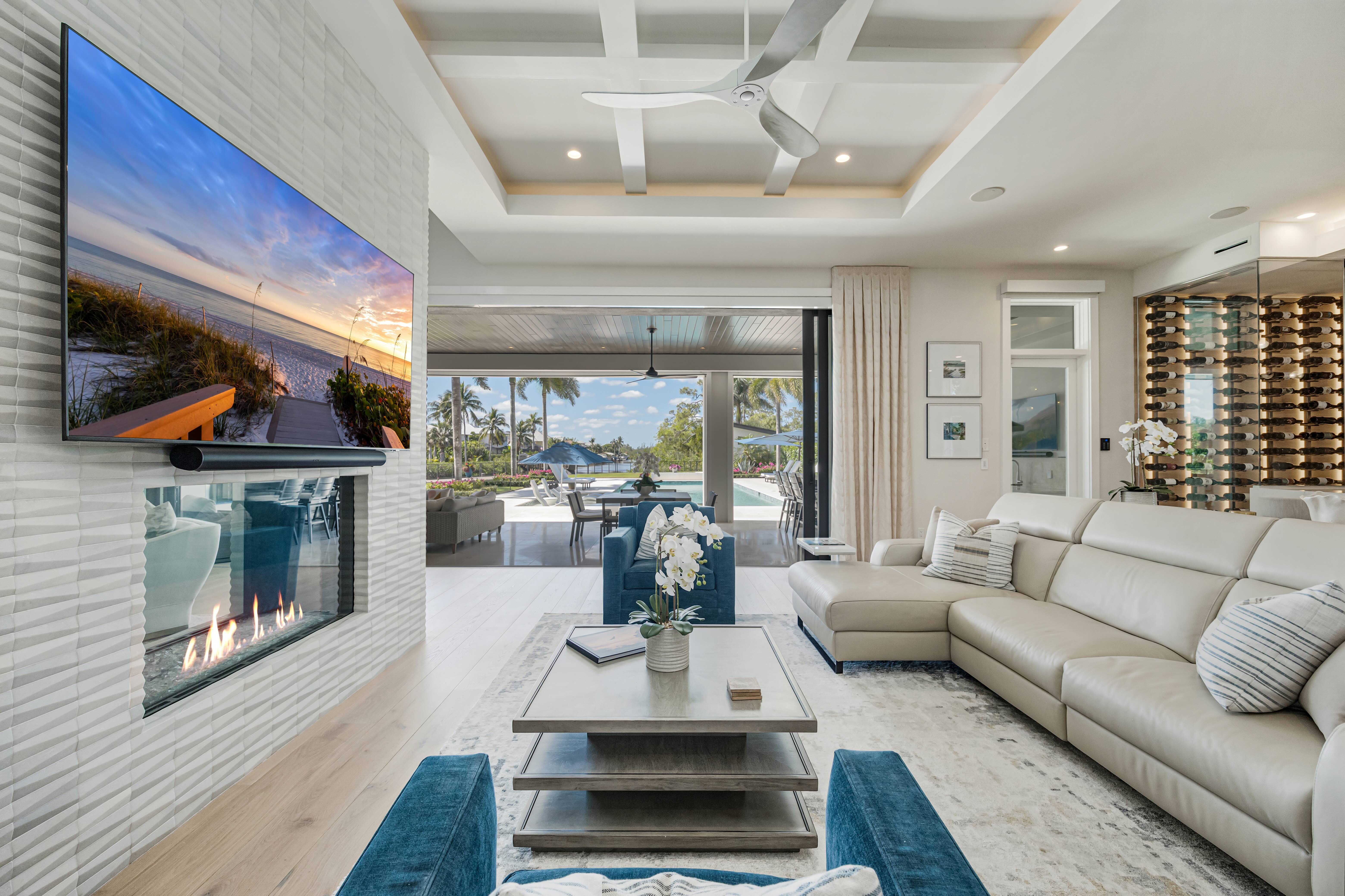 Modern living room with beige leather sectional, blue velvet chairs, a fireplace below a wall-mounted TV, and large sliding doors opening to a patio and pool area.