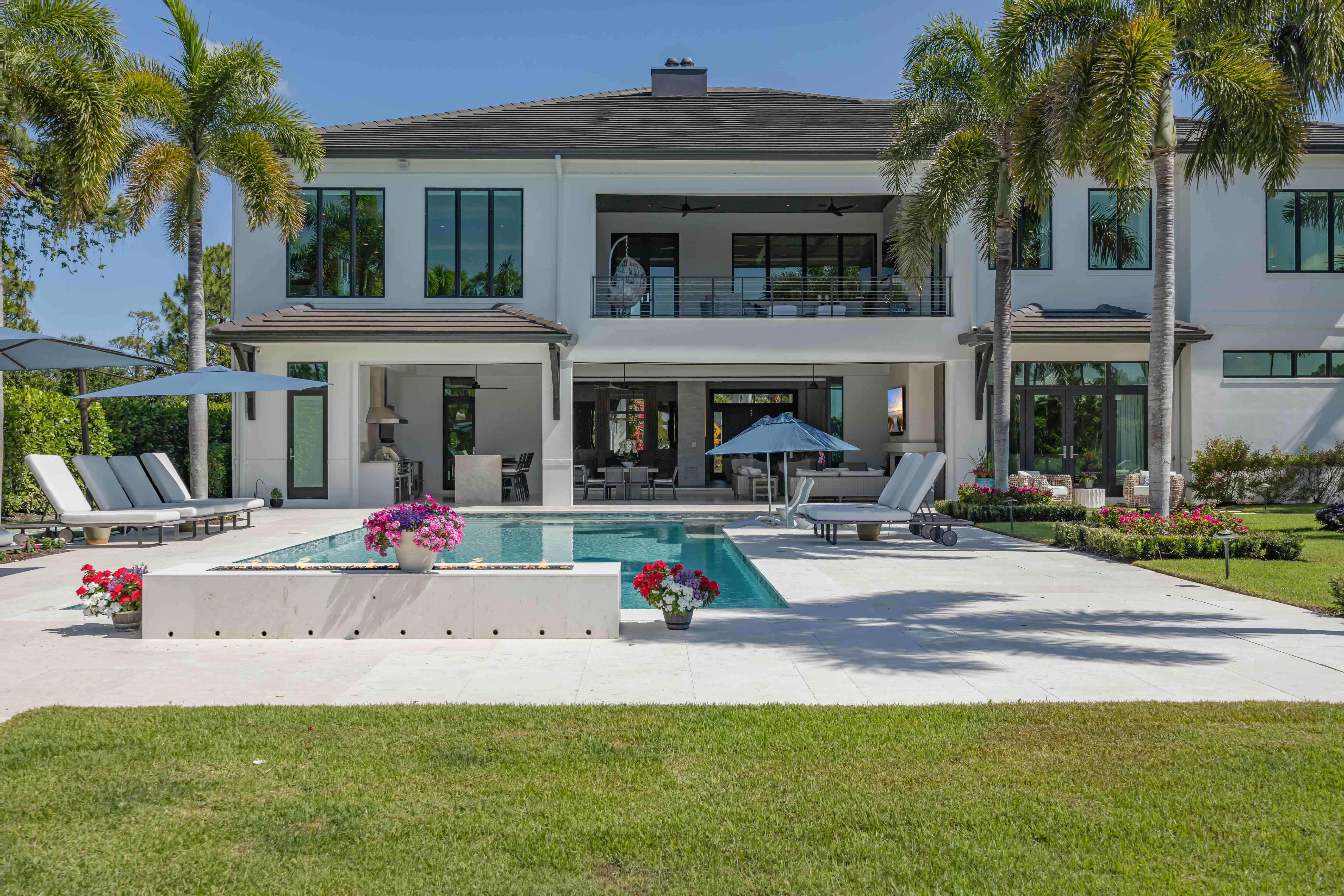 Modern two-story house with large windows, a pool, sun loungers, umbrellas, palm trees, and a well-kept lawn.