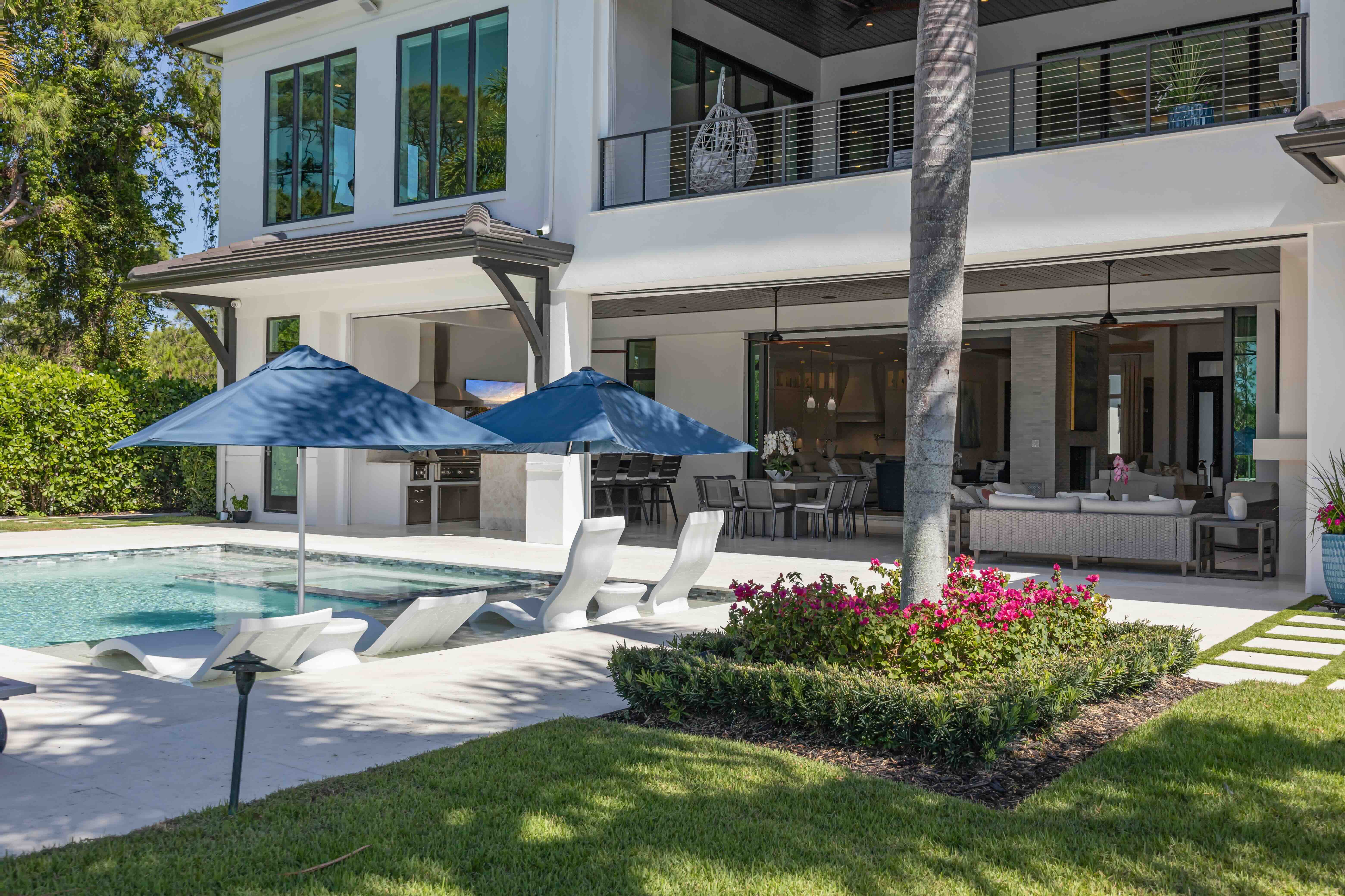 Modern two-story house with a poolside patio featuring white lounge chairs under blue umbrellas and a garden with pink flowers around a palm tree.