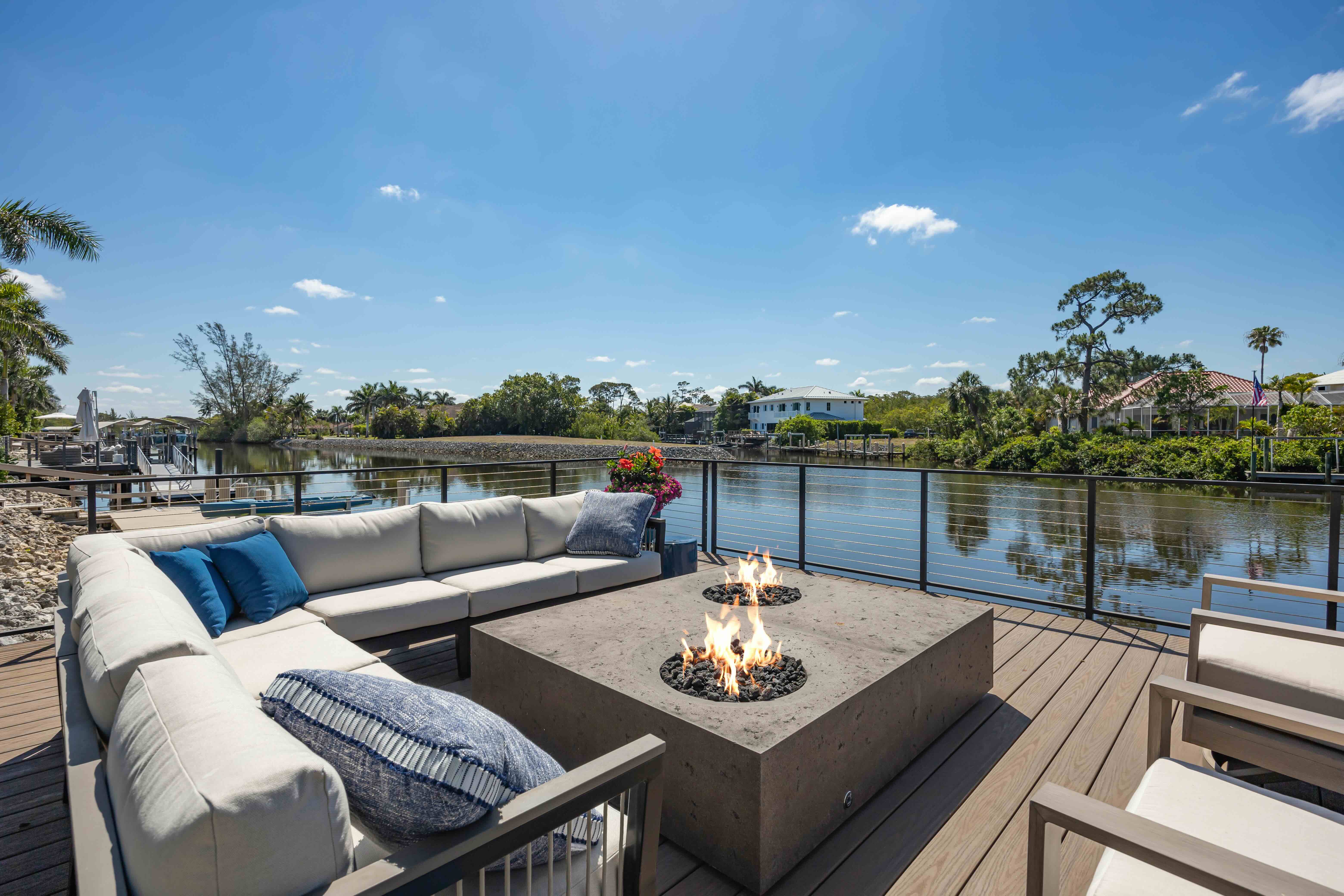 Outdoor patio with beige sectional sofa, fire pit table, and chairs overlooking a calm waterfront with houses and palm trees under a clear blue sky.
