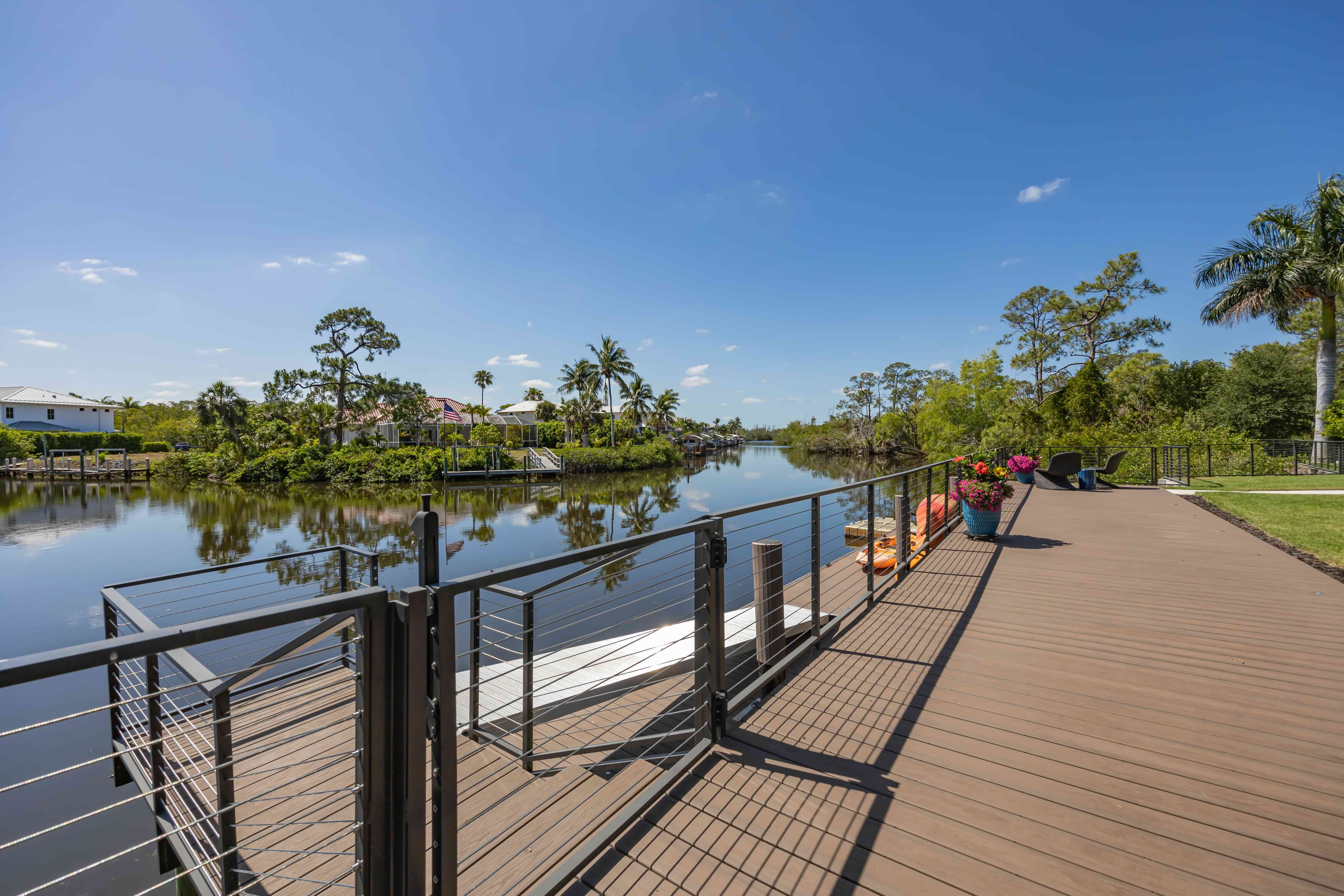 Wide wooden deck with metal railing overlooking calm river, decorated with potted flowers and outdoor chairs.