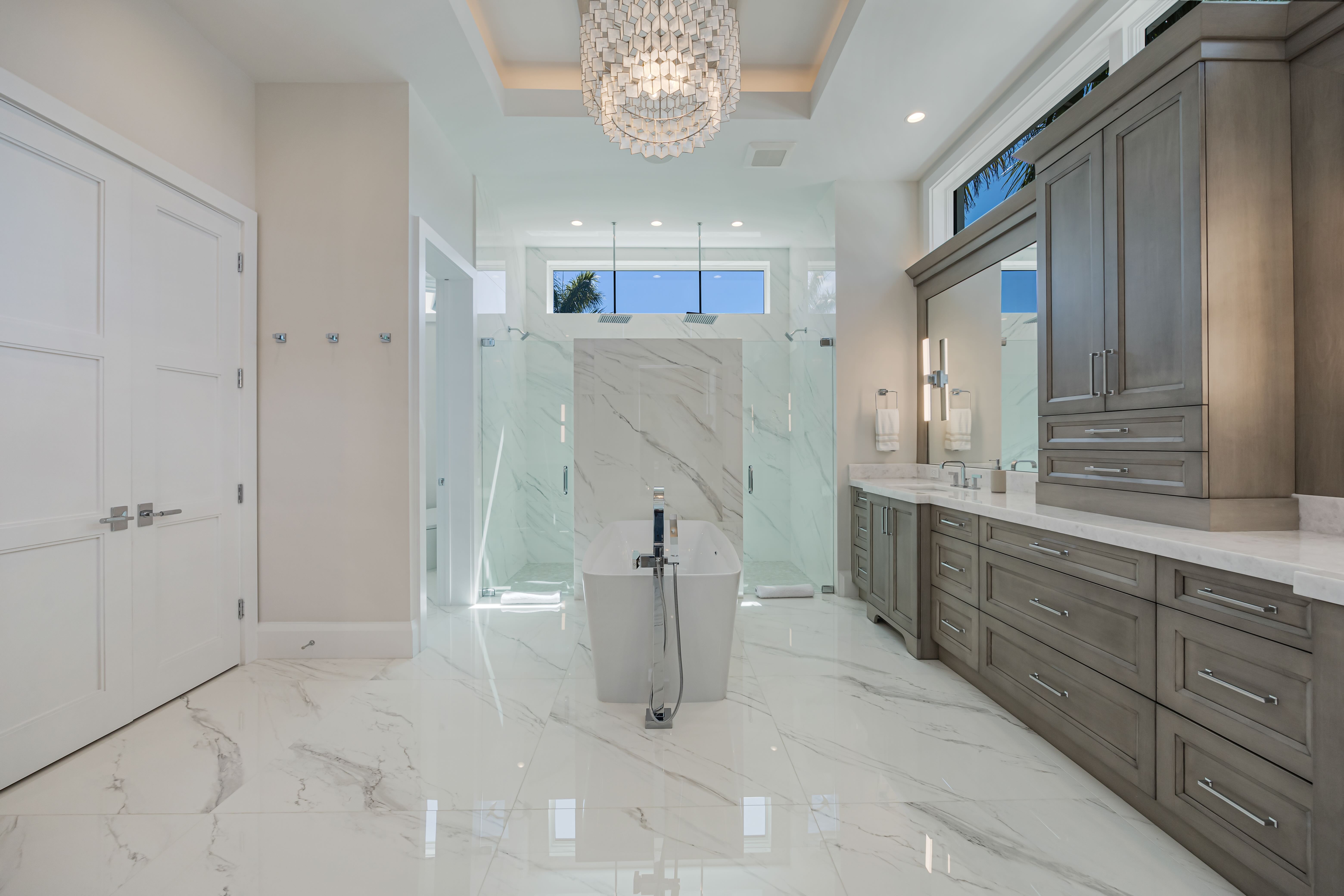 Modern bathroom with a freestanding tub, marble floors and walls, double glass shower, large wooden vanity, and a chandelier.