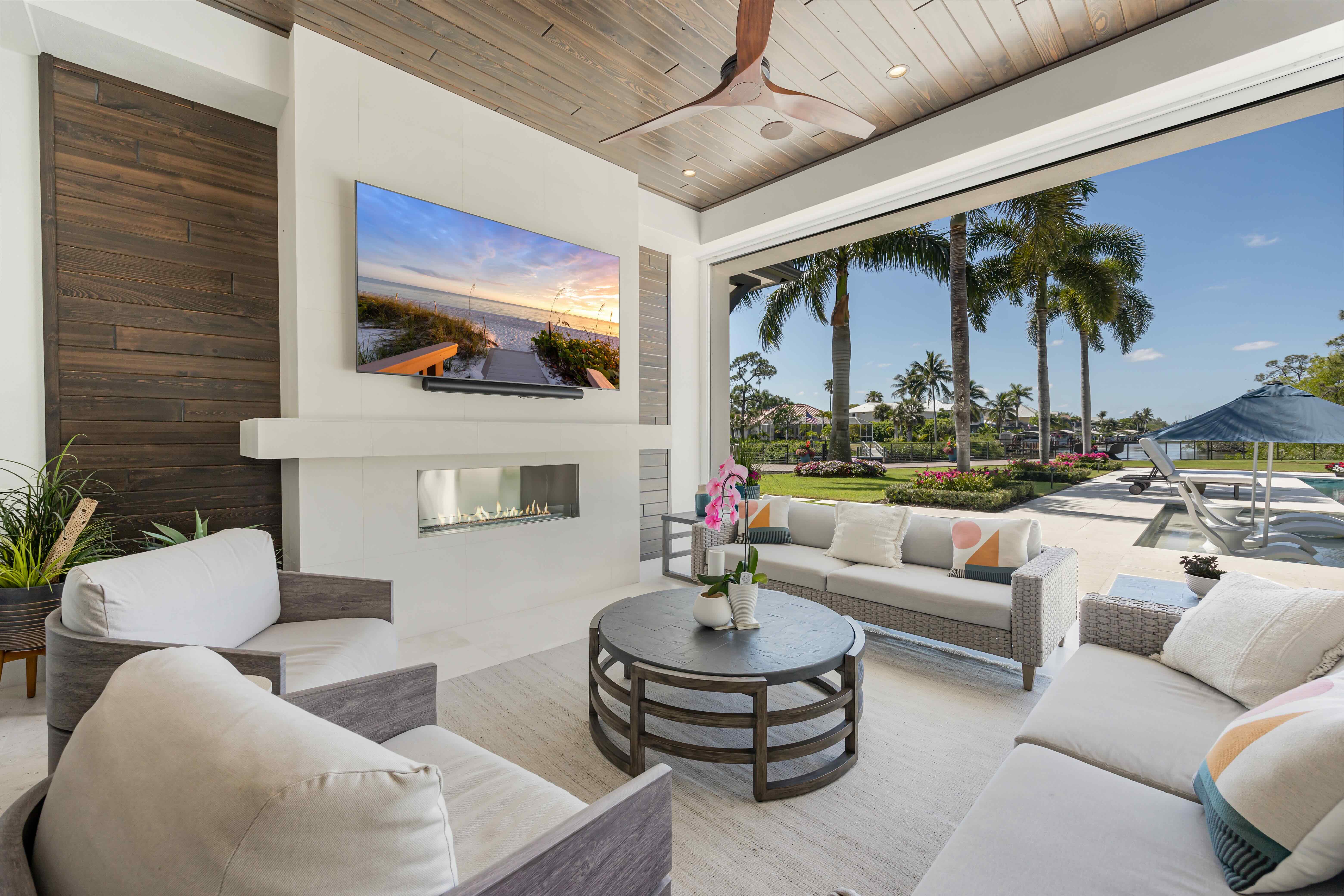 Covered patio with modern seating, round coffee table, wall-mounted TV, fireplace, and pool area overlooking palm trees and garden.