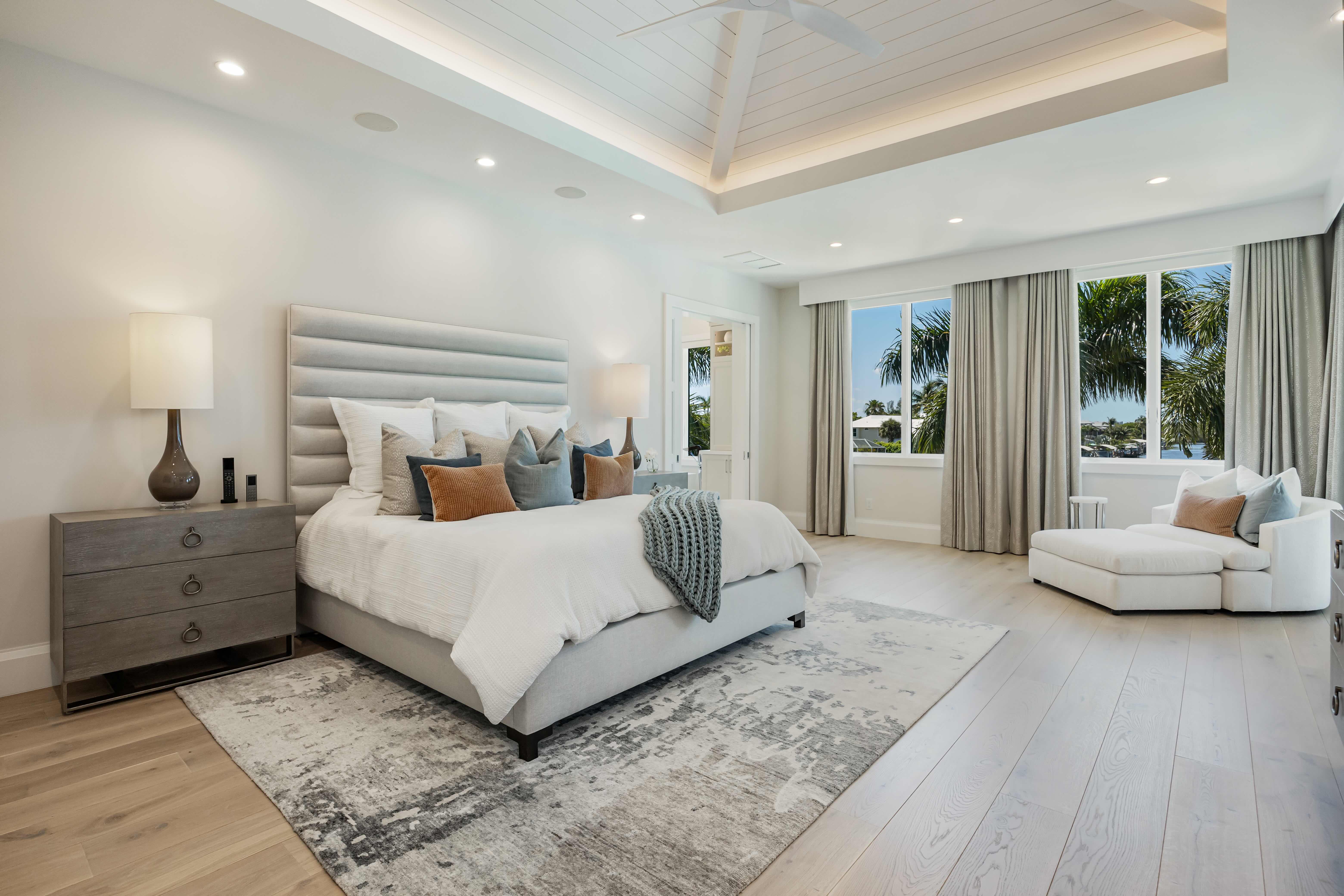 Modern bedroom with a large upholstered bed, multiple pillows, bedside tables with lamps, a gray area rug, light wood flooring, and a chaise lounge near windows with curtains showing palm trees outside.