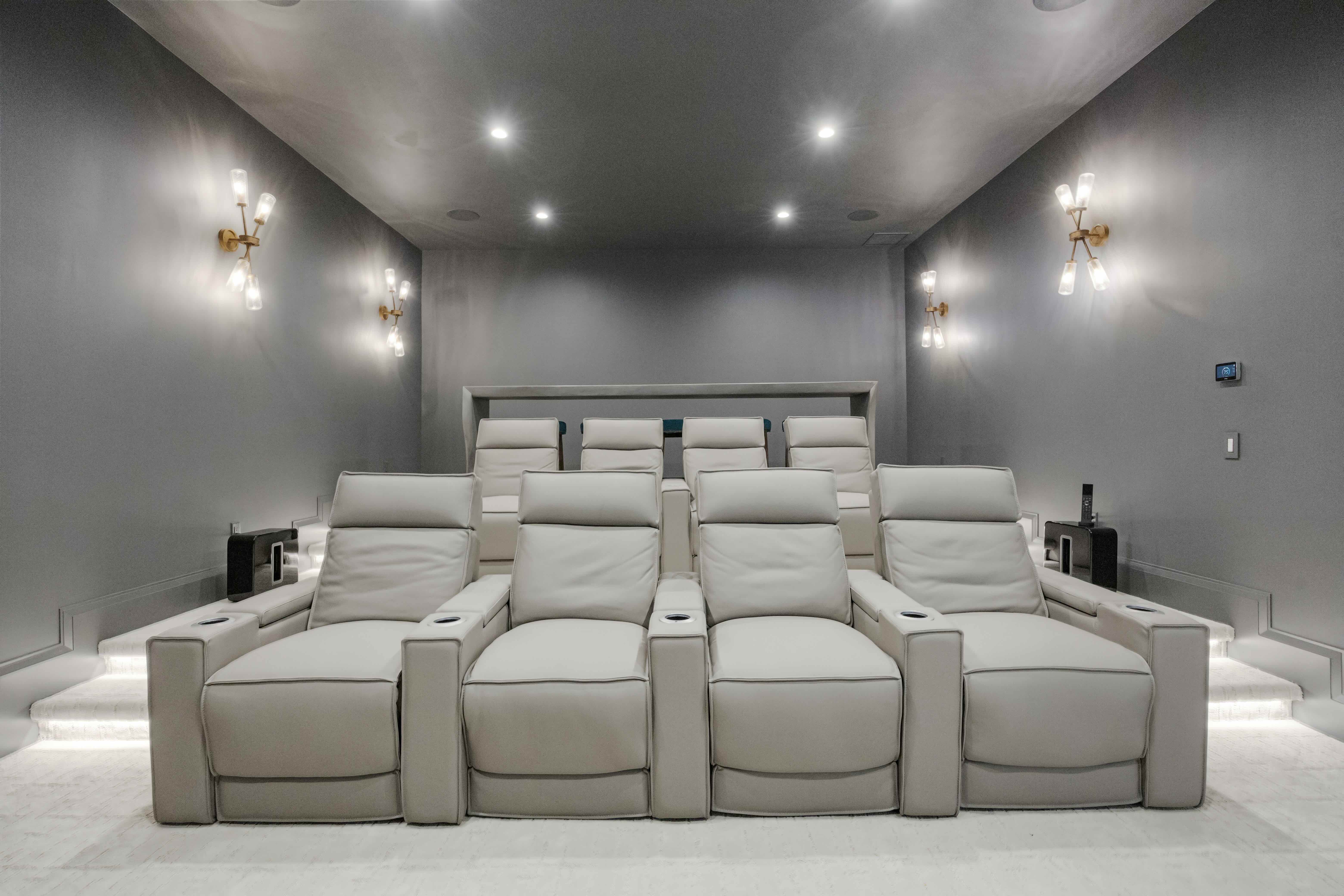 Modern home theater room with two rows of gray reclining chairs, gray walls, and wall-mounted lights.