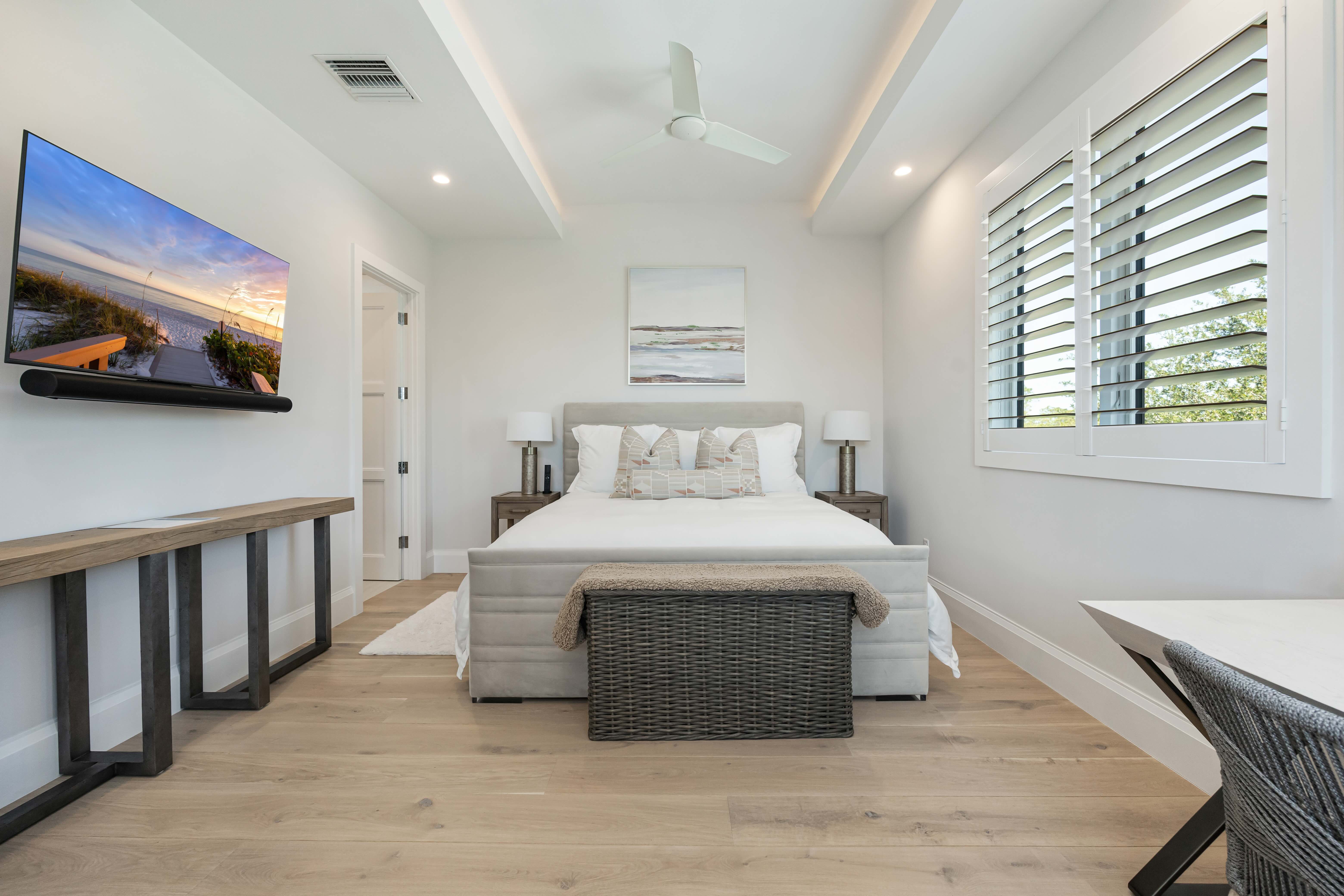 Modern bedroom with a white bed, two bedside lamps, a wicker bench, wall-mounted TV, and large window blinds.