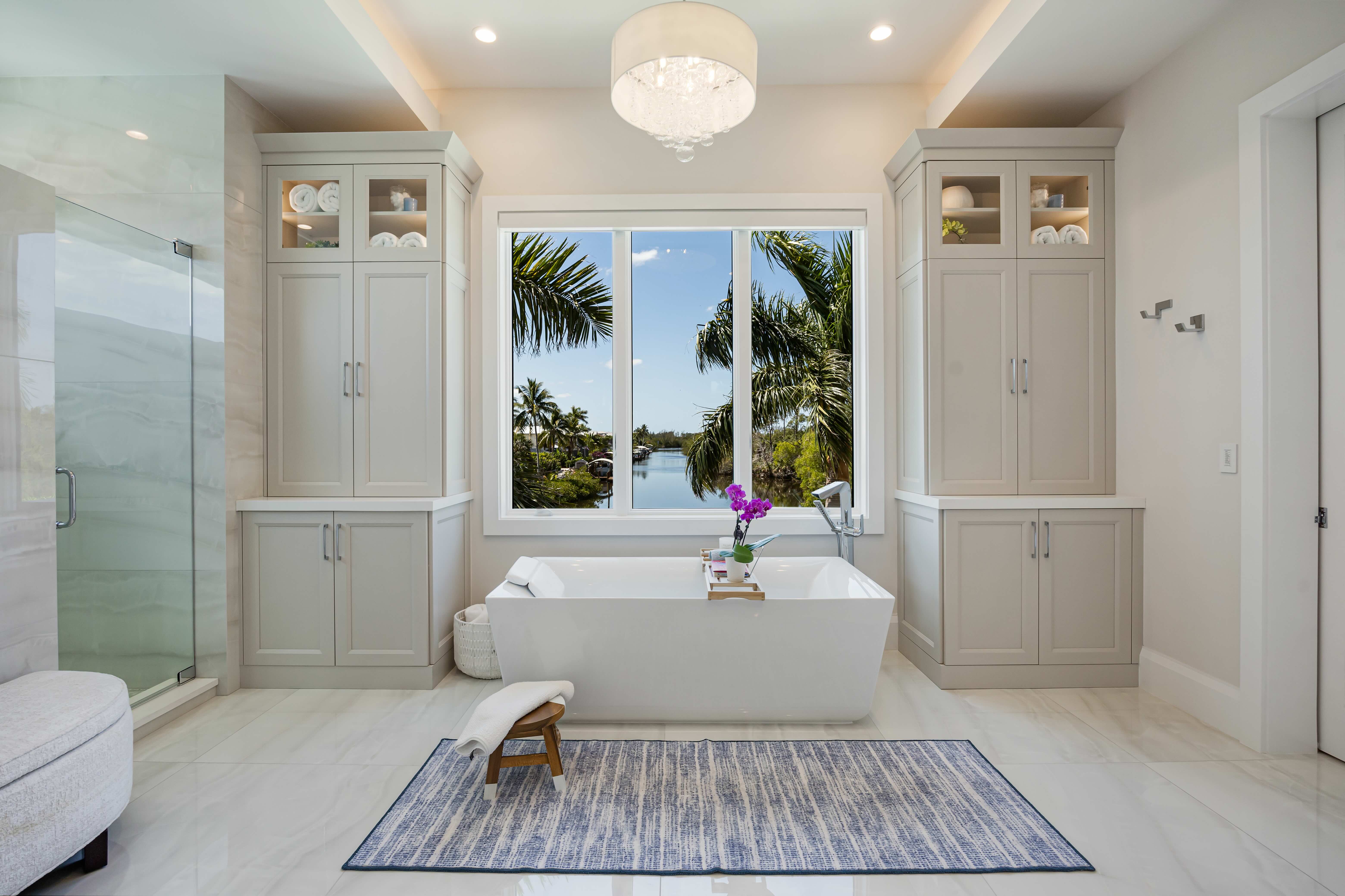 Modern bathroom with a white freestanding bathtub centered under a large window showing palm trees and water outside, flanked by built-in cabinets and a blue patterned rug on the floor.