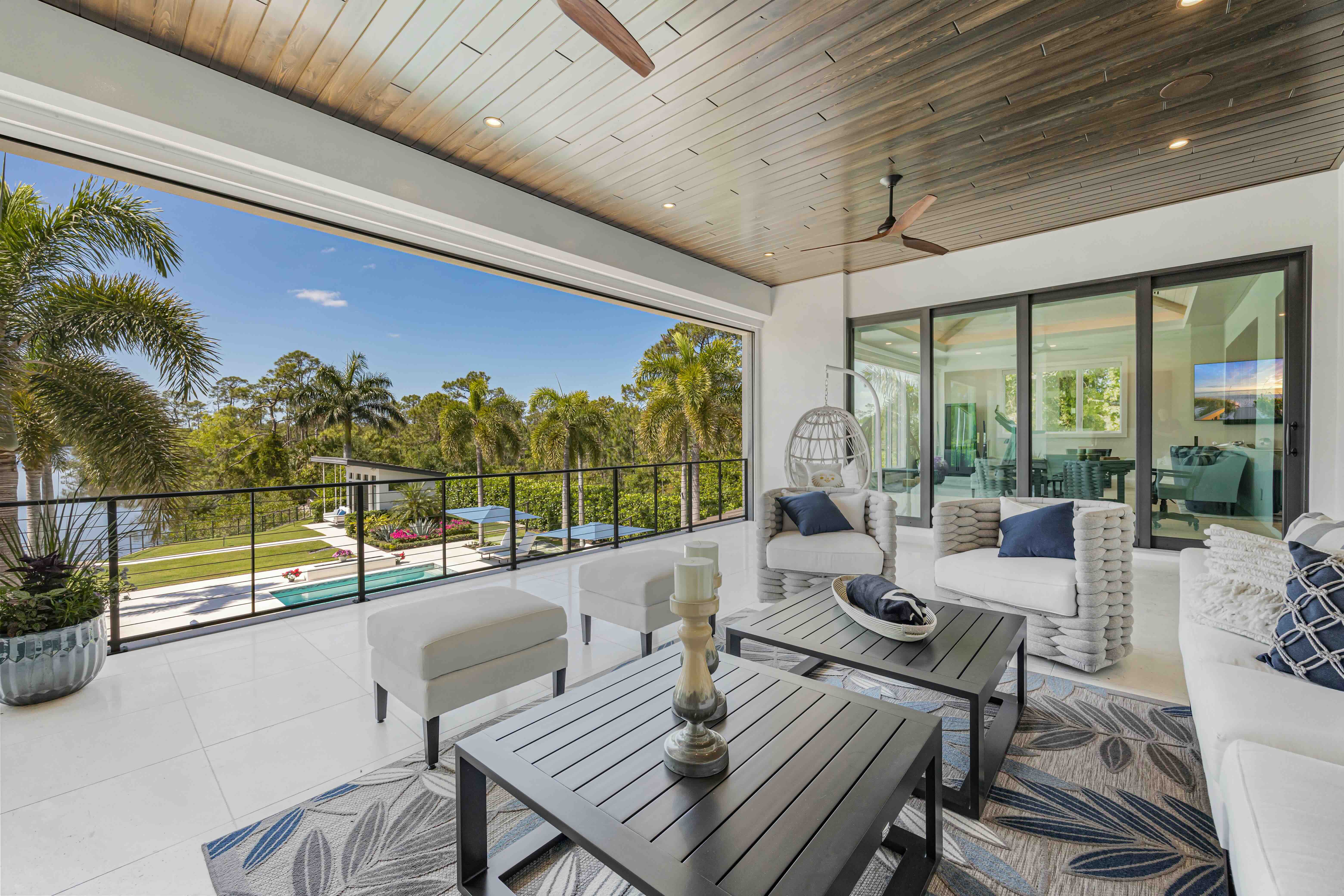 Modern covered patio with gray armchairs, black coffee tables, patterned rug, and view of palm trees and swimming pool.