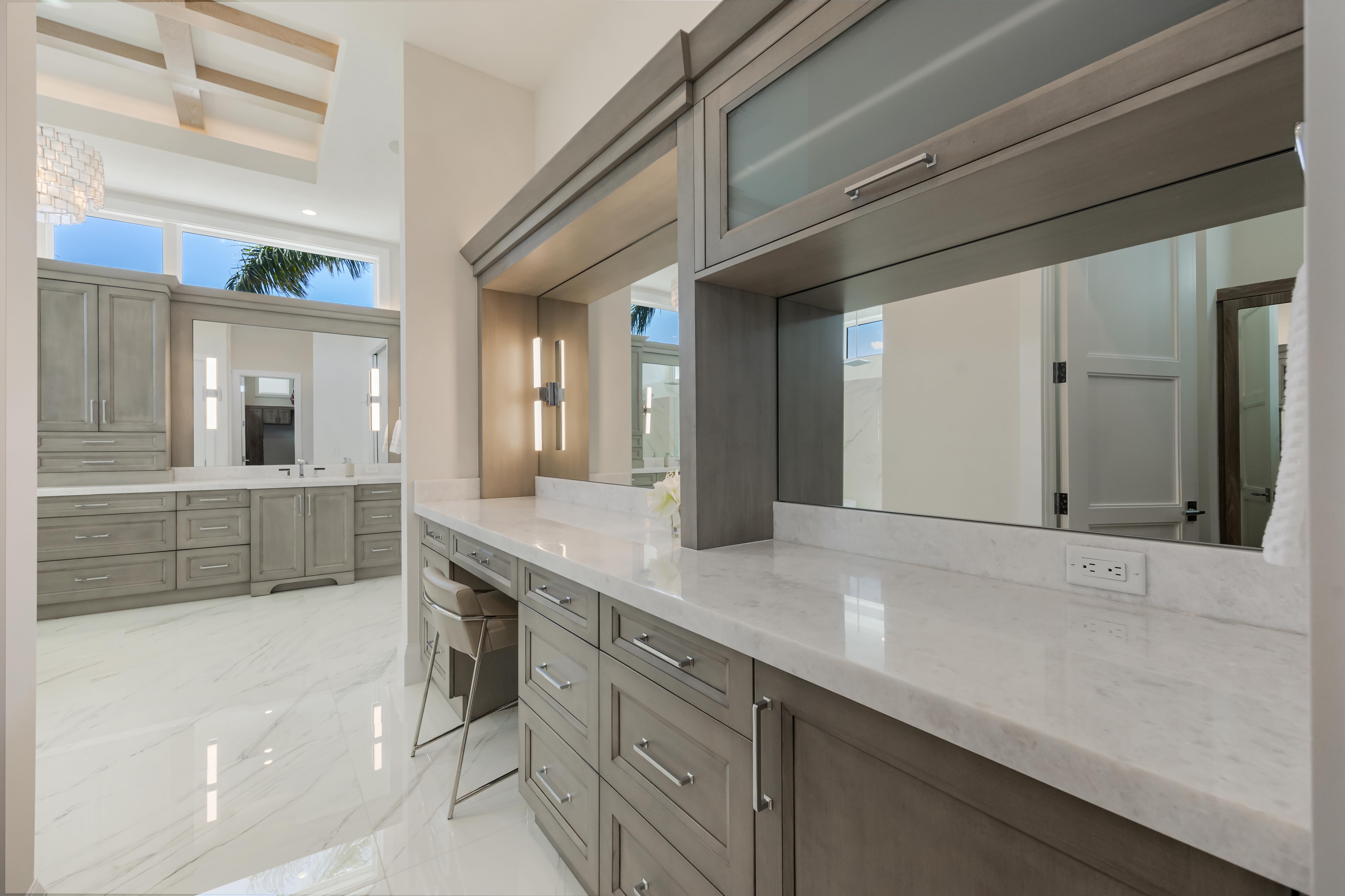 Modern bathroom vanity with gray cabinets, white marble countertop, large mirrors, and a beige cushioned stool.