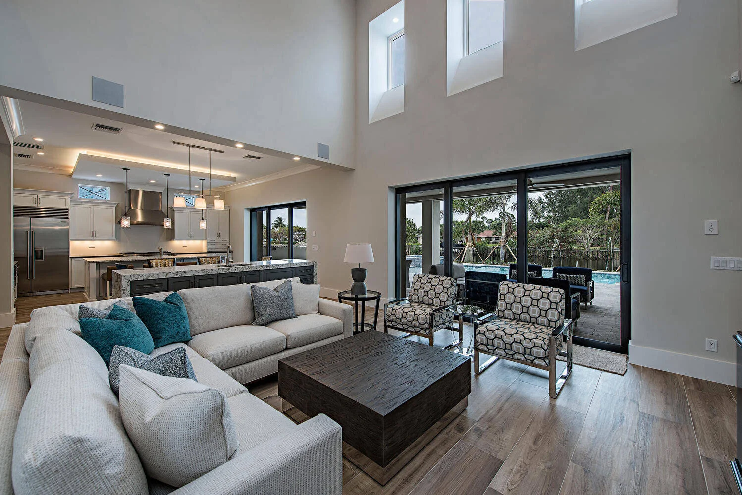 Modern living room with a large sectional sofa, patterned armchairs, dark wooden coffee table, and sliding glass doors leading to an outdoor pool area.