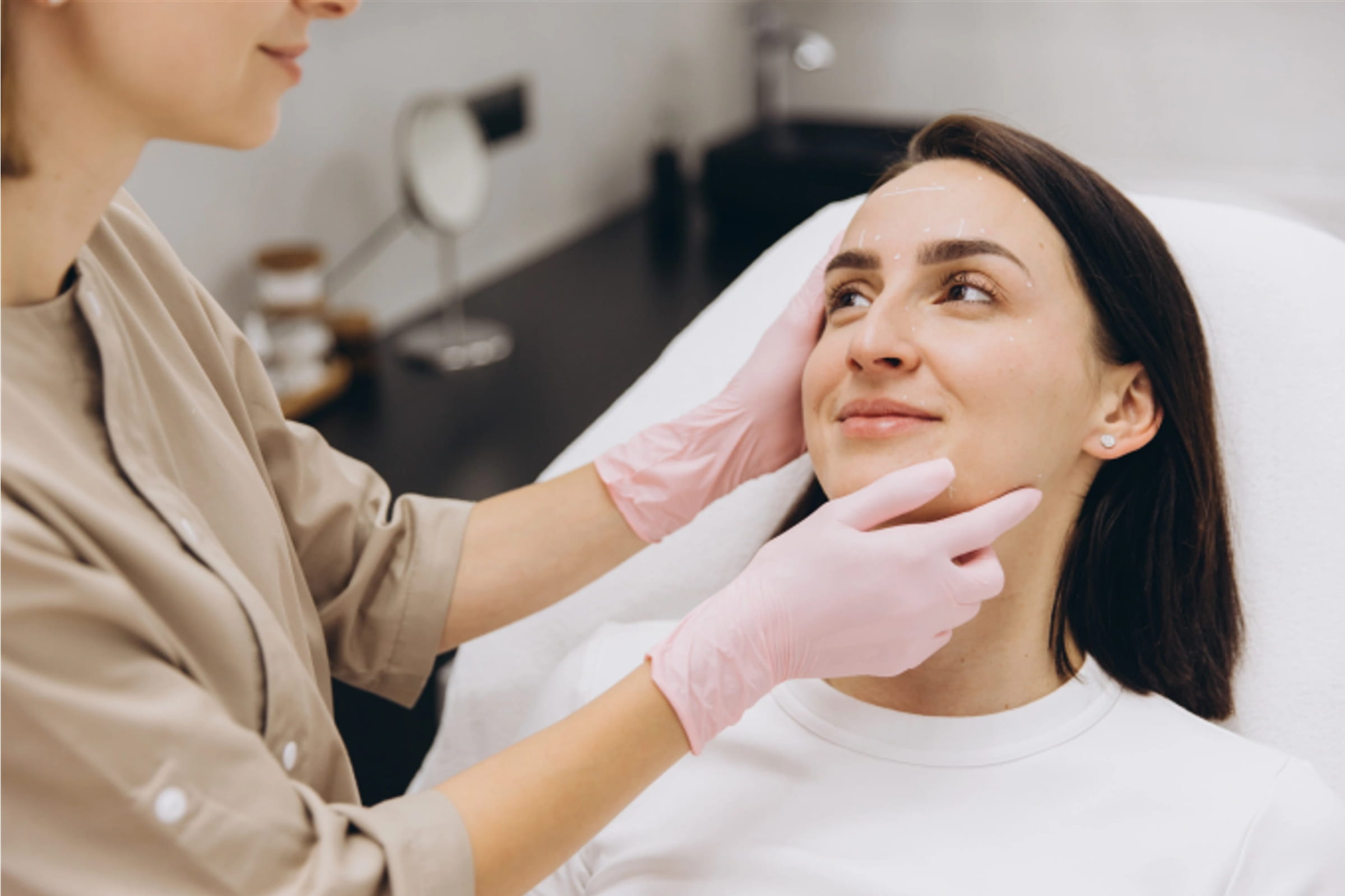 A professional aesthetician in pink gloves evaluating a smiling woman's facial skin during a dermaplaning consultation at a medical spa.