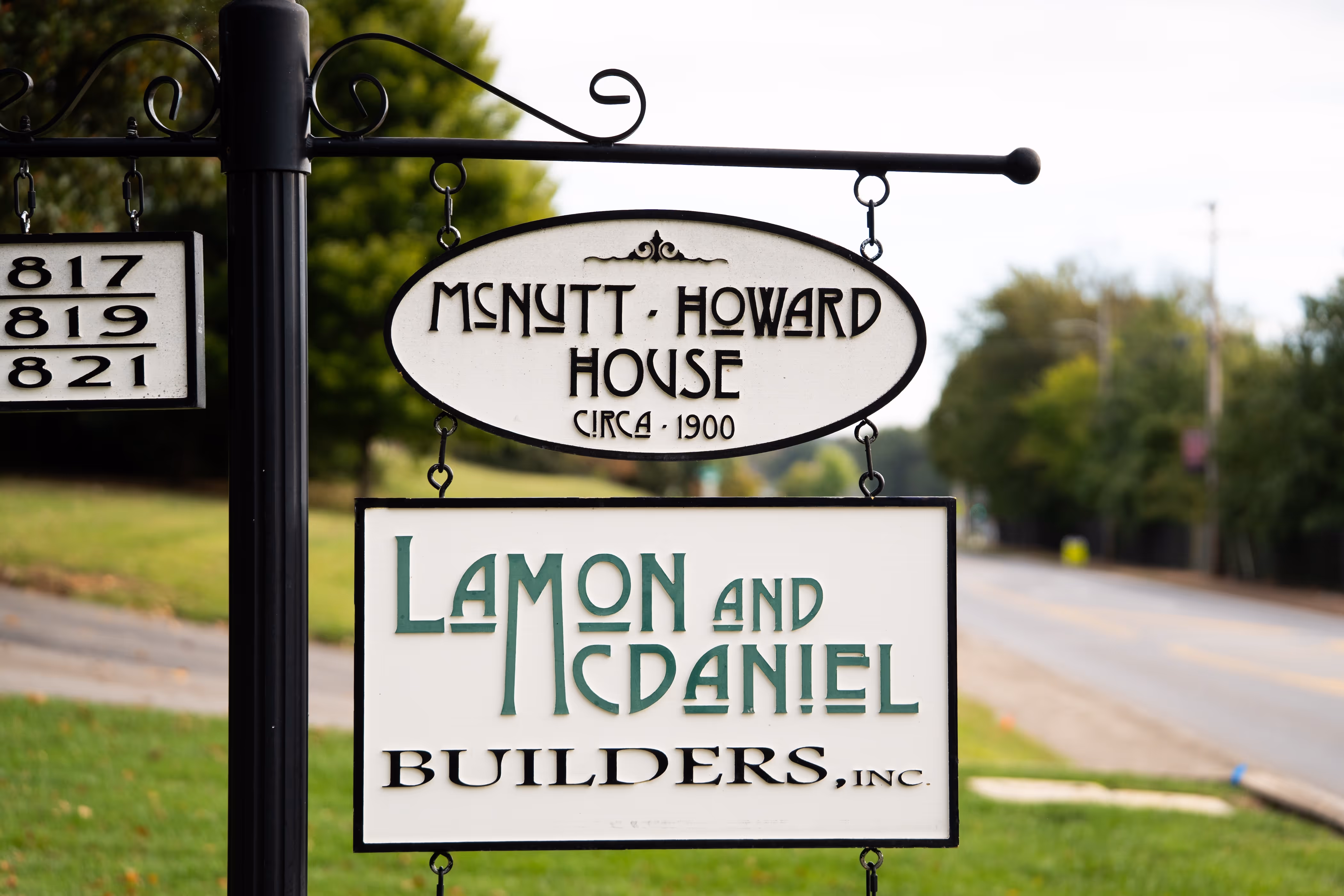 Street sign for McNutt-Howard House circa 1900 and Lamon and McDaniel Builders, Inc. along a roadside with green grass and trees.