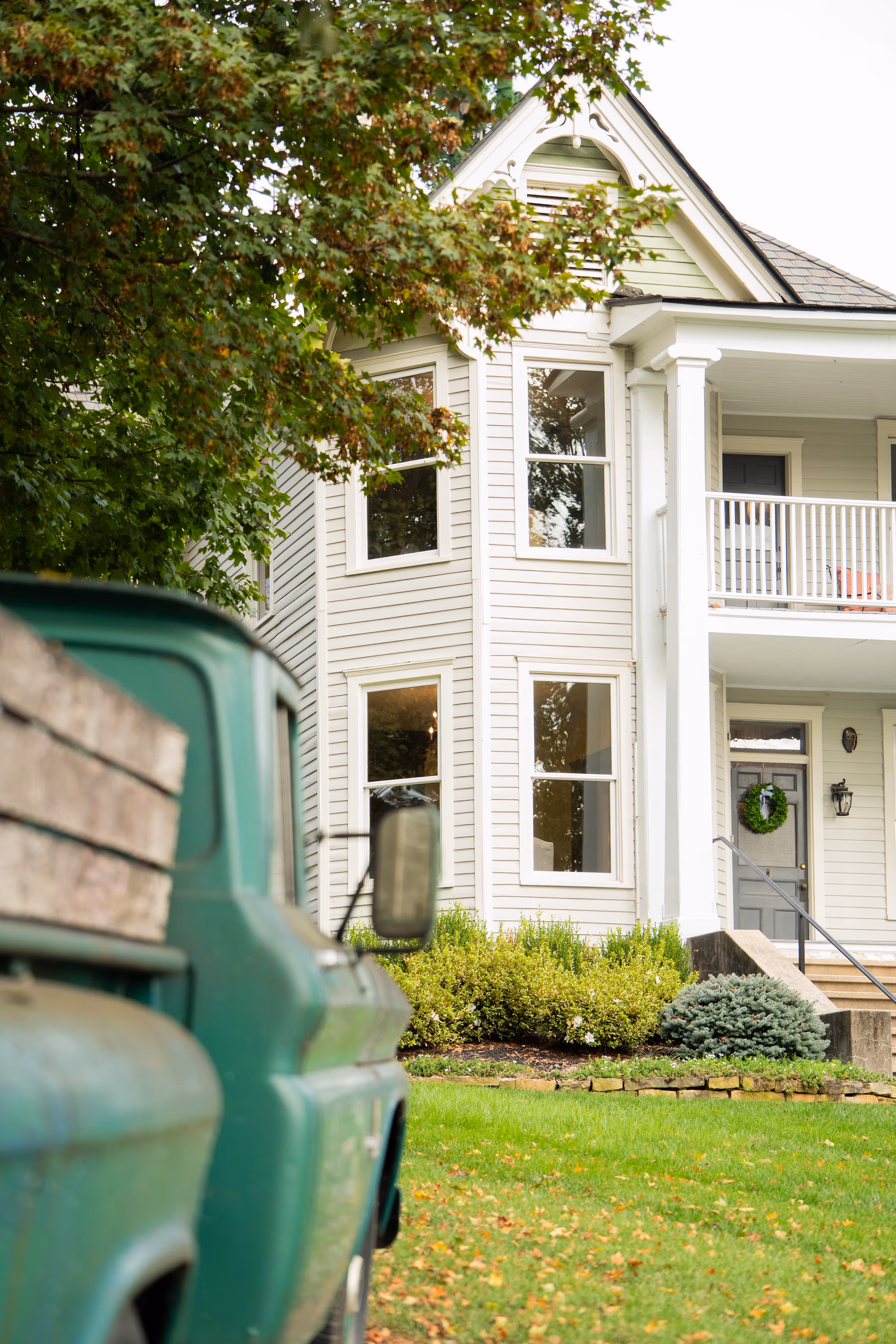 Green vintage pickup truck parked near a white two-story house with a front porch and a wreath on the door surrounded by green bushes and trees.