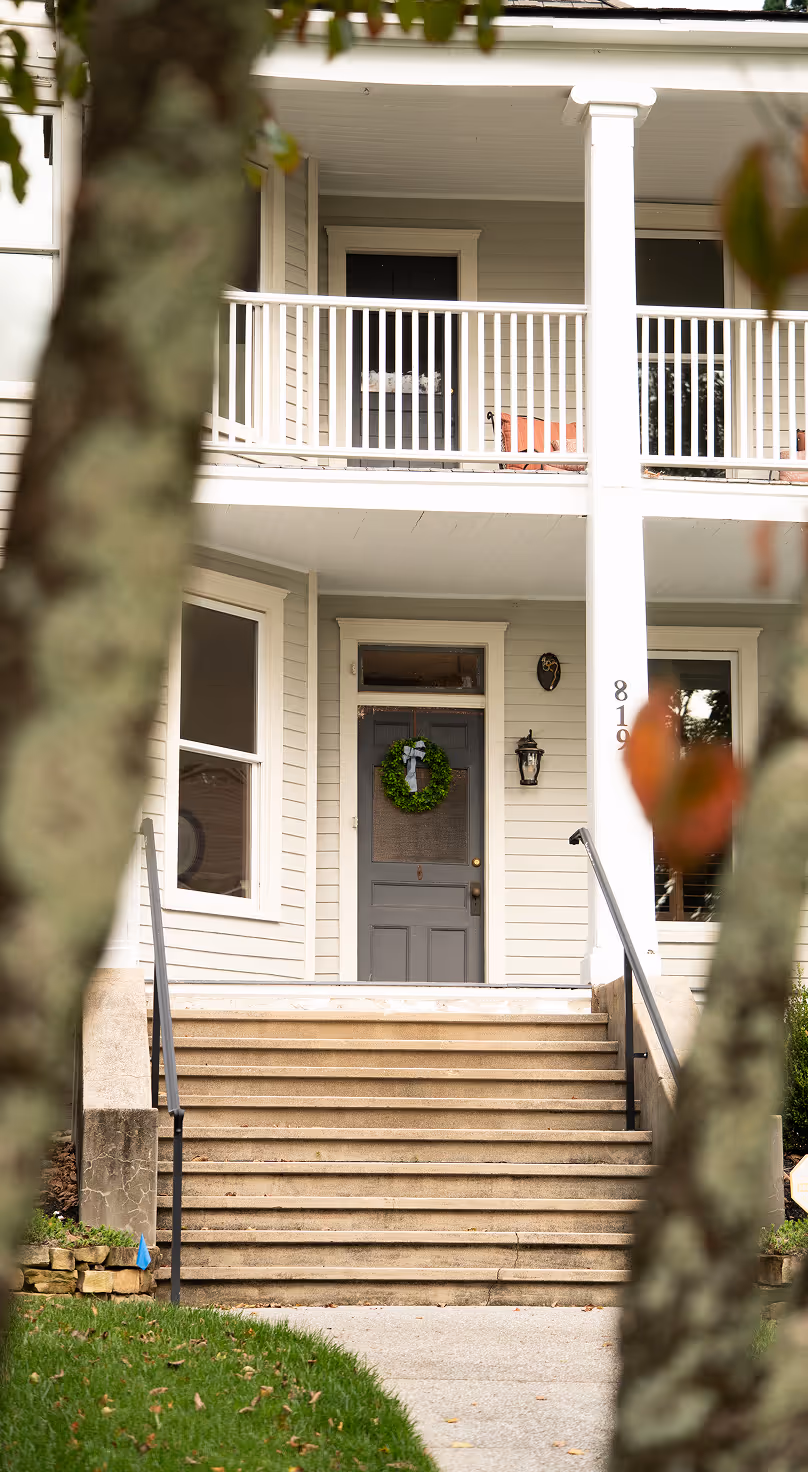 Front entrance of a white house with stone steps, a gray door decorated with a green wreath, white railing, and the house number 819 on a column.