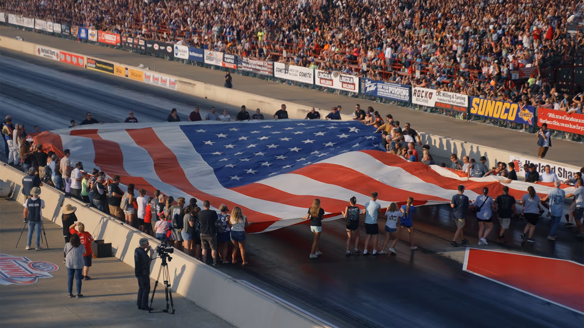 A large group of people holding and displaying a giant American flag on a race track with a crowd in the stands.