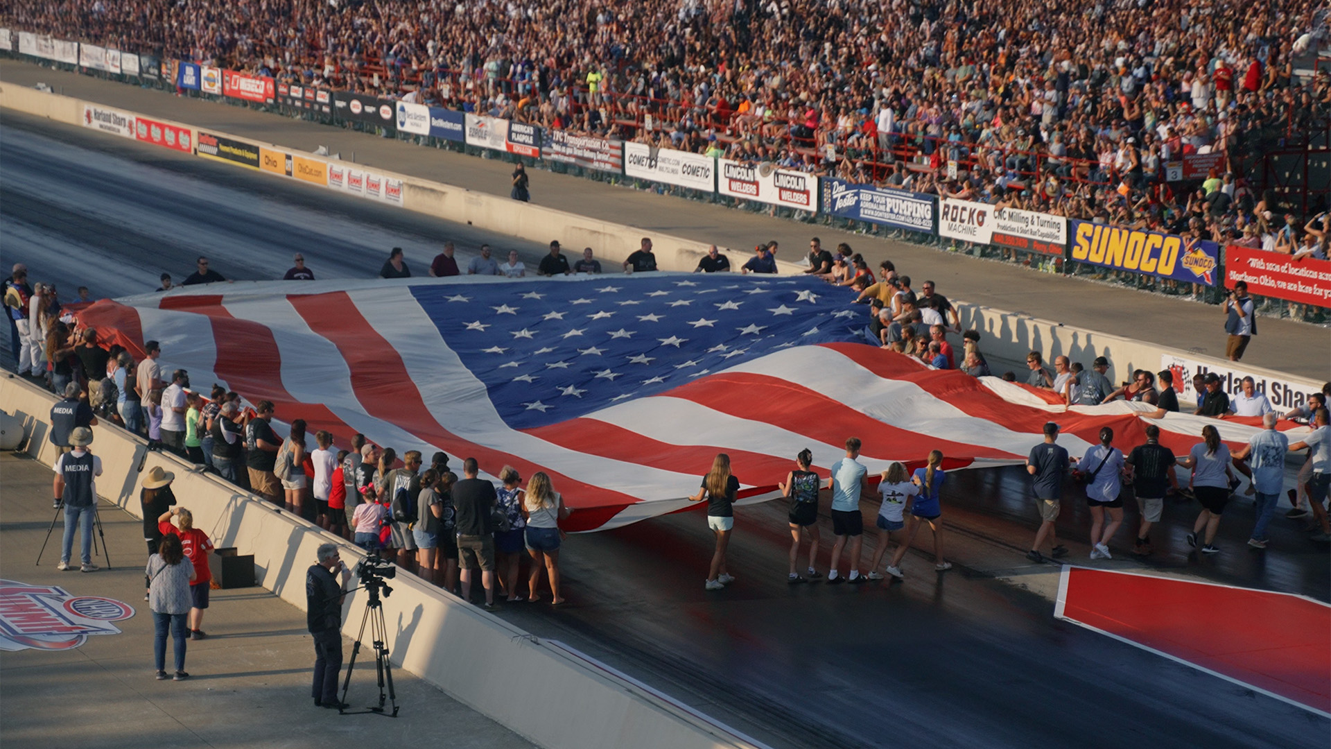 A large group of people holding and displaying a giant American flag on a race track with a crowd in the stands.