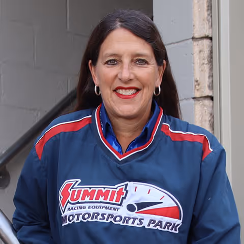 Woman with long dark hair smiling, wearing a blue Summit Racing Equipment Motorsports Park jacket with red and white accents.