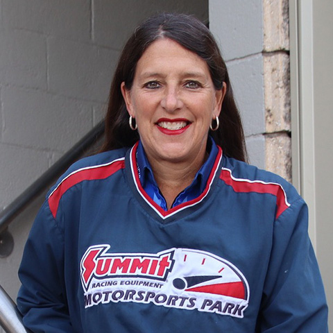 Woman with long dark hair smiling, wearing a blue Summit Racing Equipment Motorsports Park jacket with red and white accents.