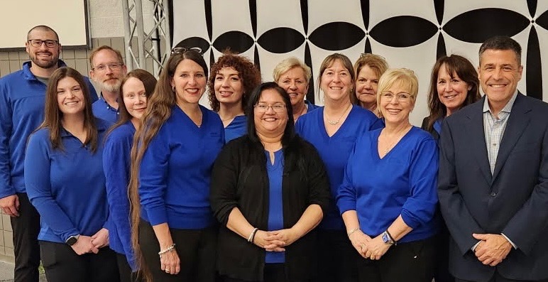 Group of twelve adults standing indoors, eleven wearing blue tops and one man in a dark suit, smiling at the camera.
