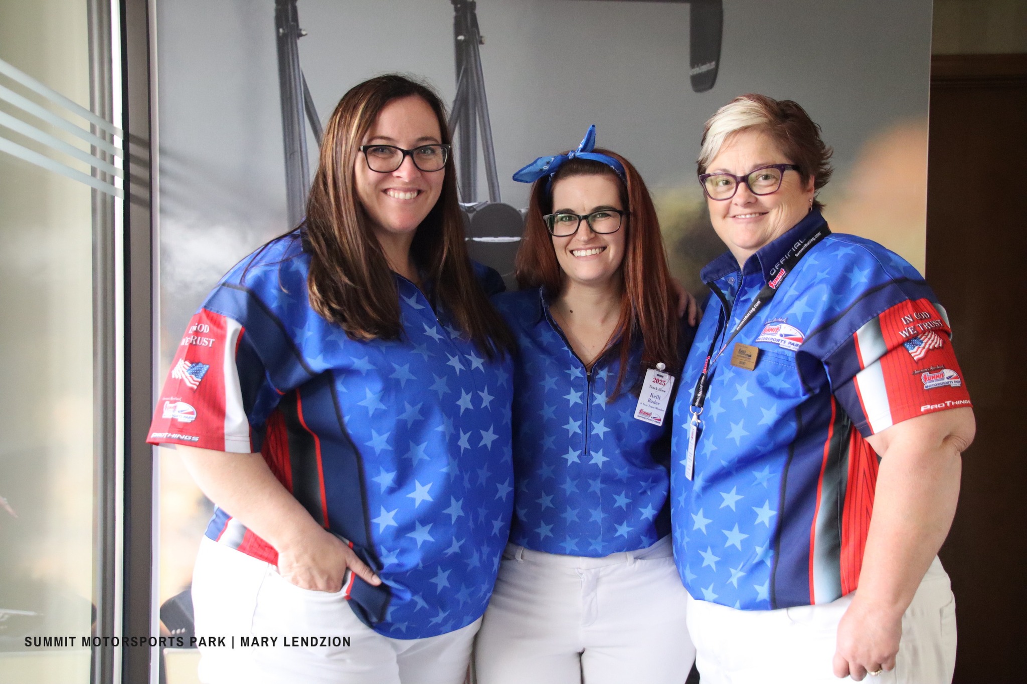 Three women standing together smiling, wearing blue star-patterned shirts and white pants, posing indoors.