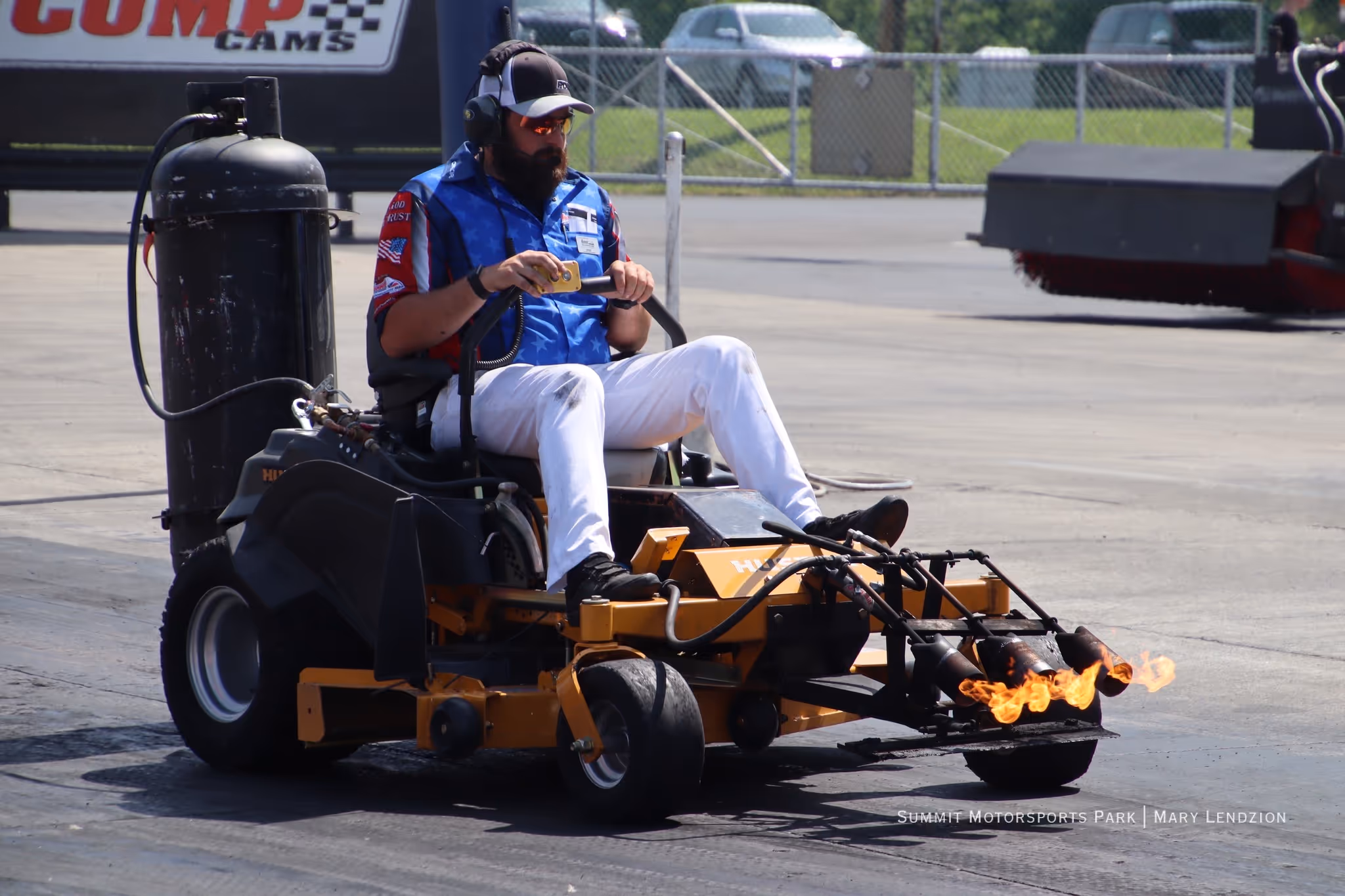Man with beard and sunglasses driving a yellow racing lawn mower with flames shooting from the front nozzles.