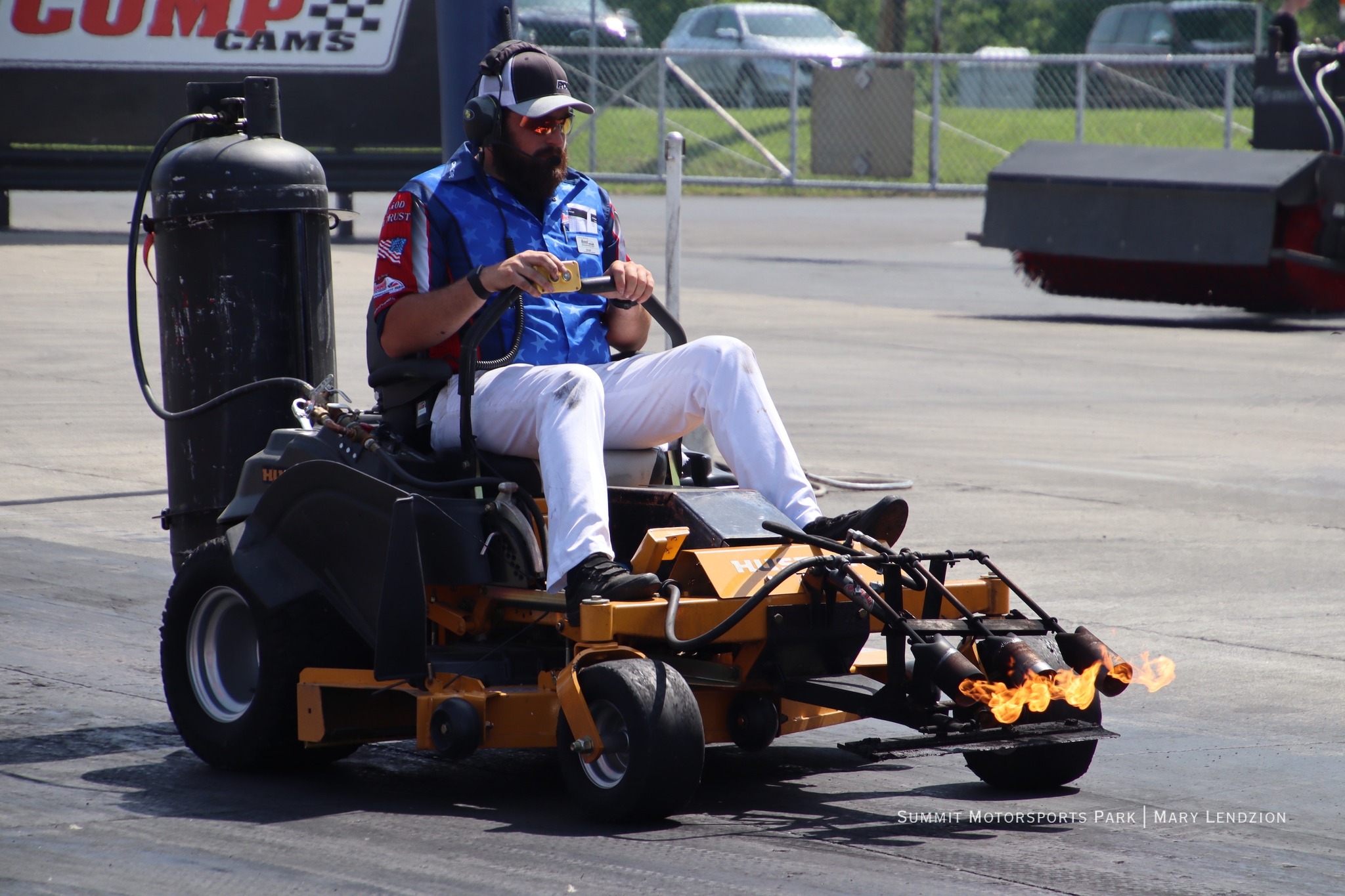 Man with beard and sunglasses driving a yellow racing lawn mower with flames shooting from the front nozzles.