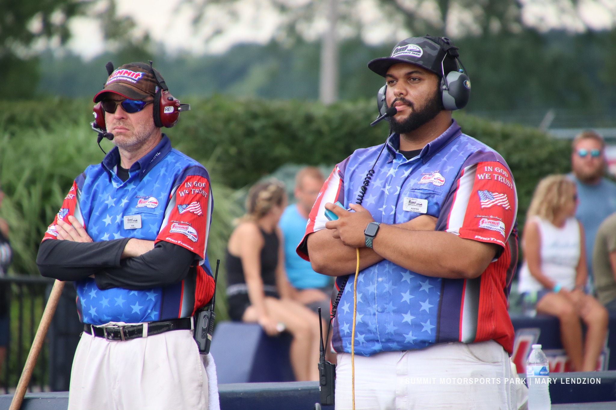 Two men wearing blue and red Summit Motorsports Park uniforms with headsets standing with arms crossed at an outdoor event.