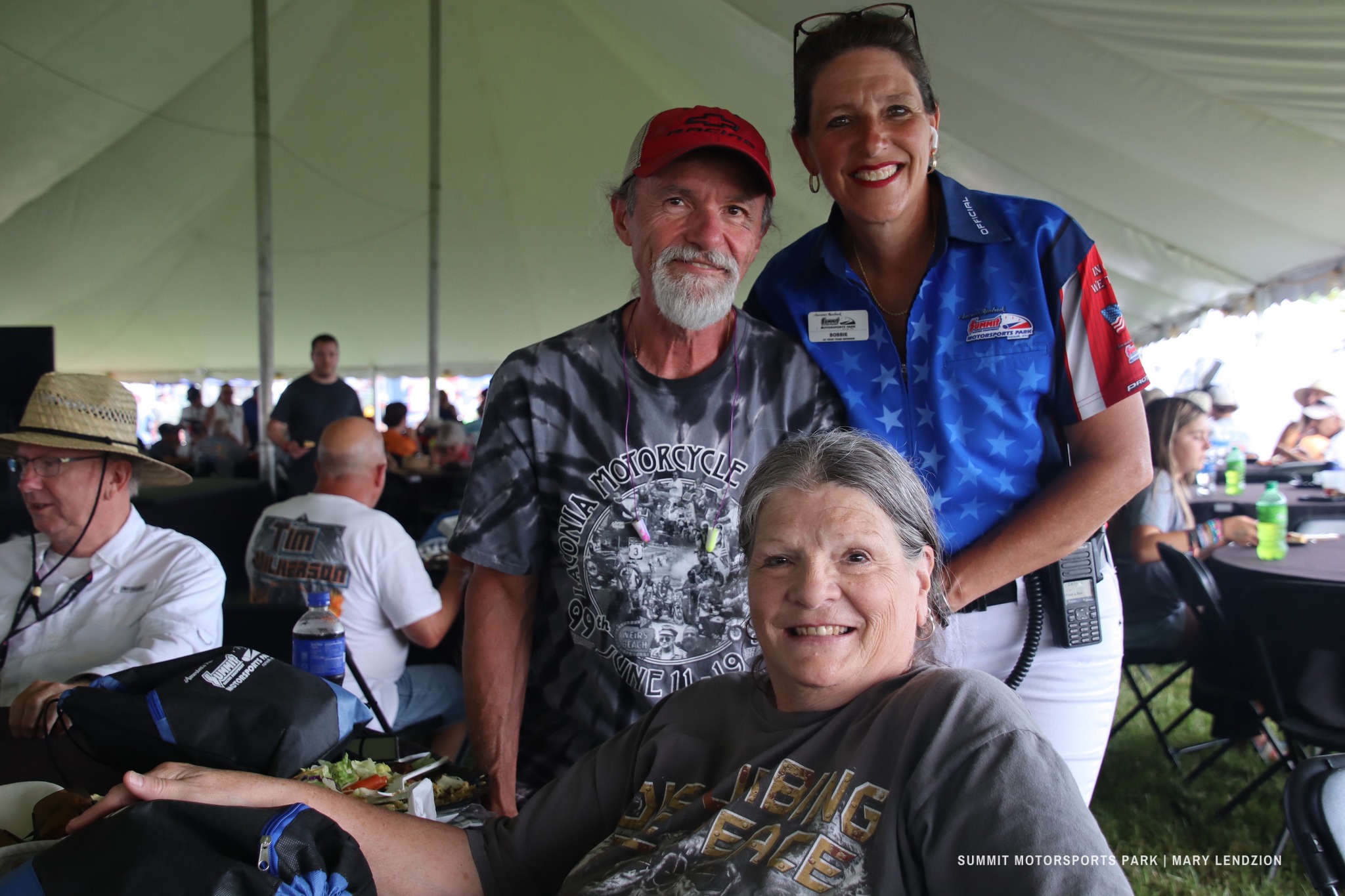 Three smiling adults posing together under a large event tent with people seated at tables in the background.
