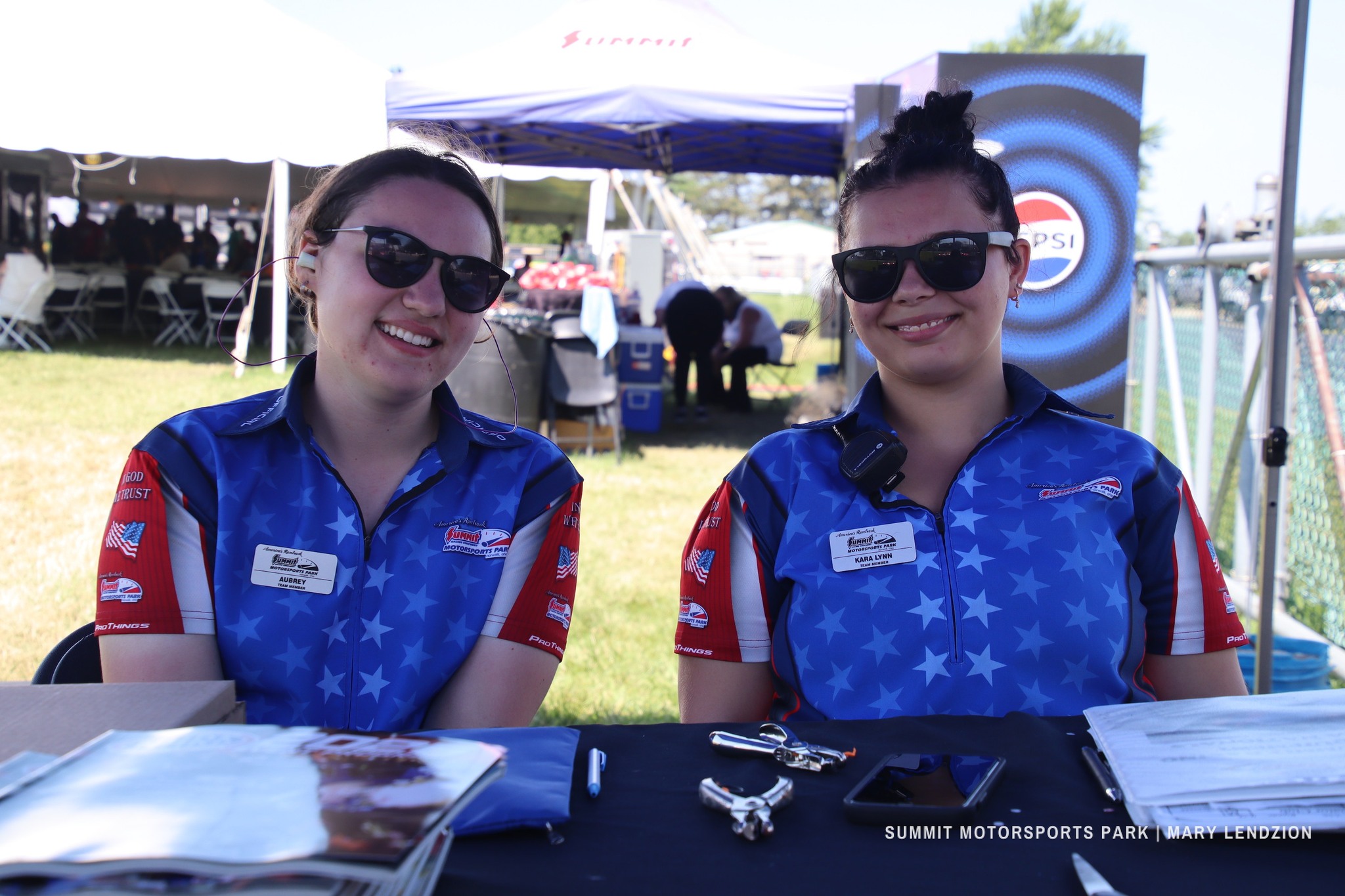Two women wearing blue Summit Motorsports Park uniforms and sunglasses sitting at a table with documents and tools outdoors.