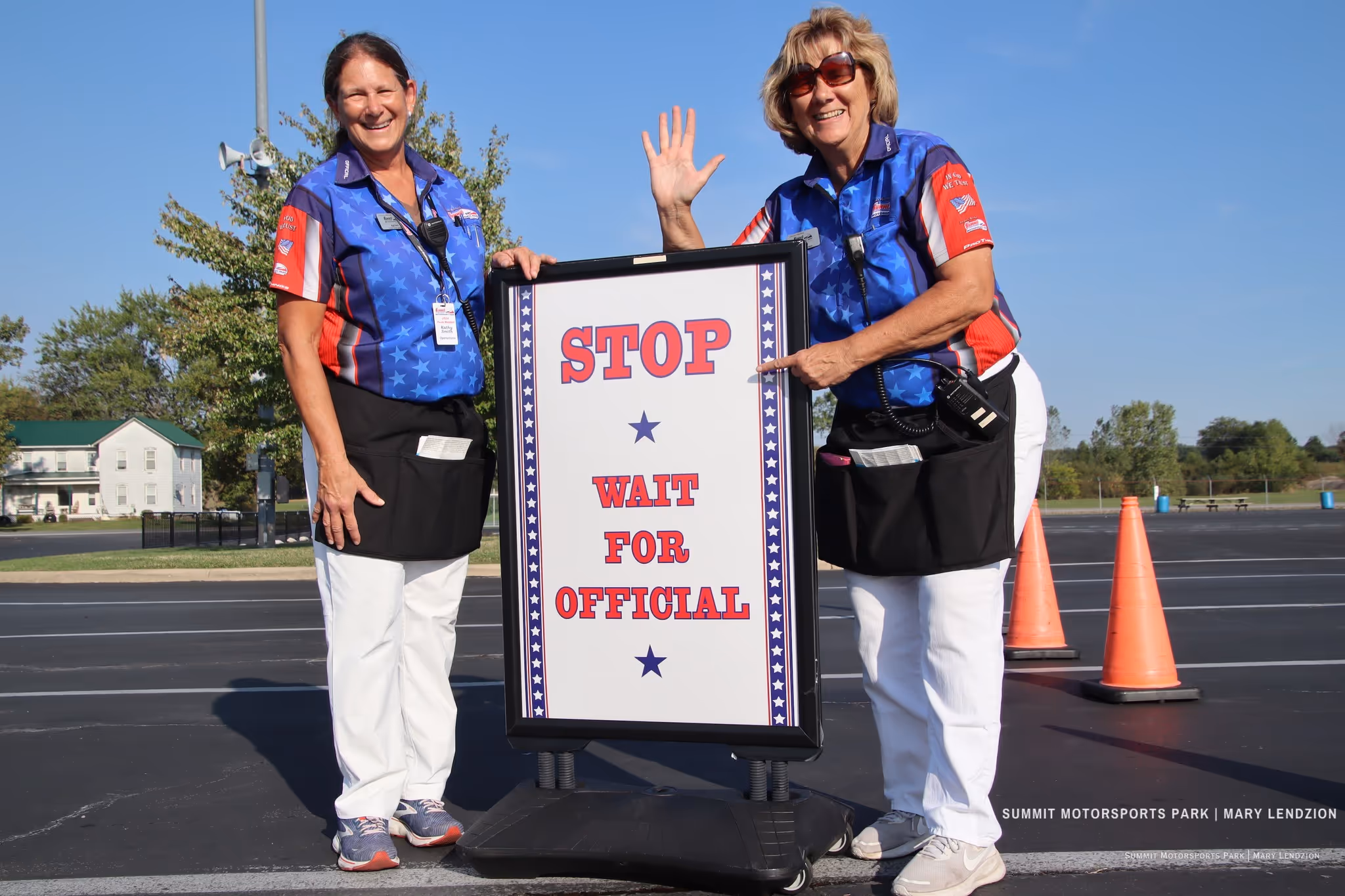 Two women in blue star-patterned shirts and white pants standing outdoors, one waving and the other pointing to a sign that reads 'STOP WAIT FOR OFFICIAL'.