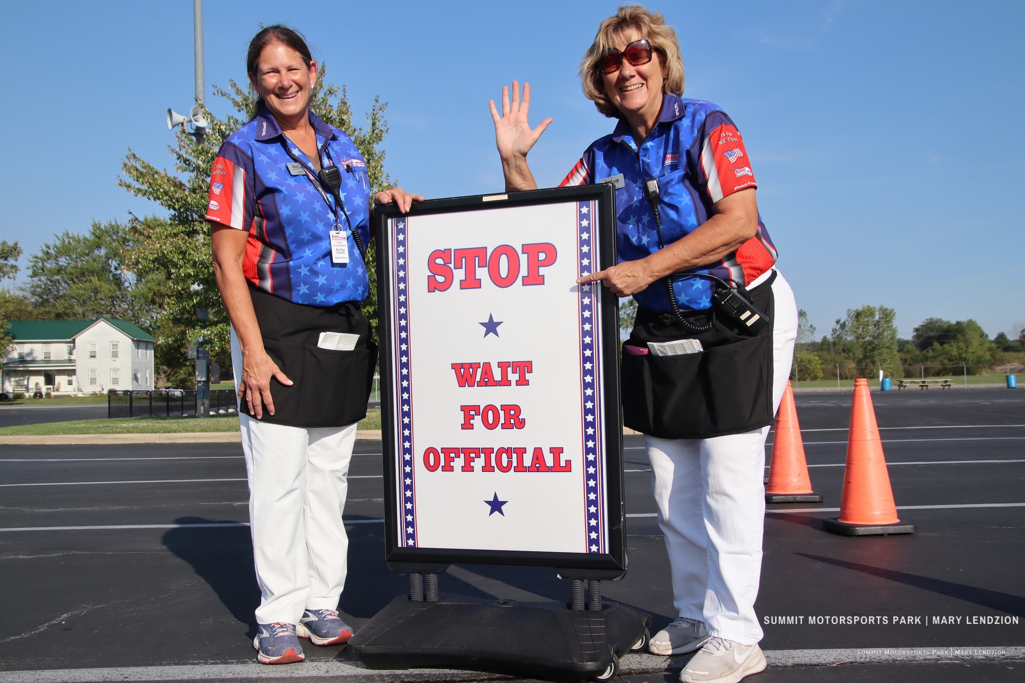Two women in blue star-patterned shirts and white pants standing outdoors, one waving and the other pointing to a sign that reads 'STOP WAIT FOR OFFICIAL'.