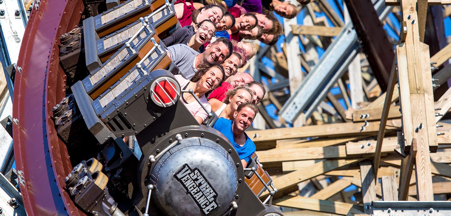 Group of people smiling and enjoying on the Steel Vengeance roller coaster with visible wooden and metal track structure.