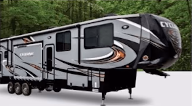 Gray and black fifth-wheel RV trailer with three axles parked on a white surface with trees in the background.