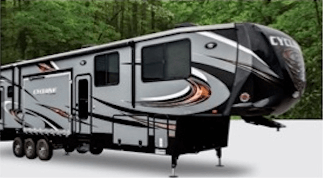 Gray and black fifth-wheel RV trailer with three axles parked on a white surface with trees in the background.