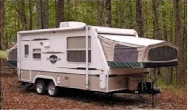 White and beige expandable camper trailer parked in a wooded area with trees and fallen leaves.