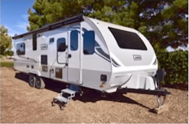 White travel trailer parked on dirt with steps extended, surrounded by trees.