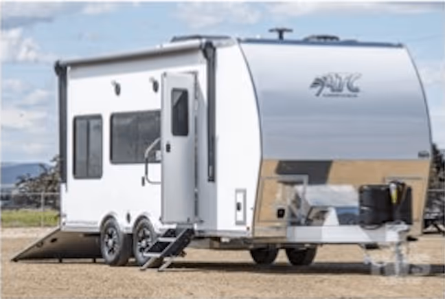 White toy hauler RV with dual axles, side entry door, and rear ramp door open, parked on a dirt surface under a partly cloudy sky.