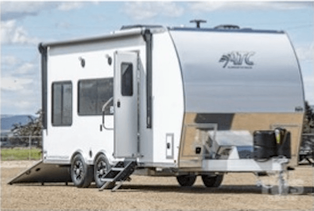 White toy hauler RV with dual axles, side entry door, and rear ramp door open, parked on a dirt surface under a partly cloudy sky.
