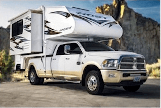White pickup truck with a large camper attached, parked on a paved surface with rocky cliffs in the background.