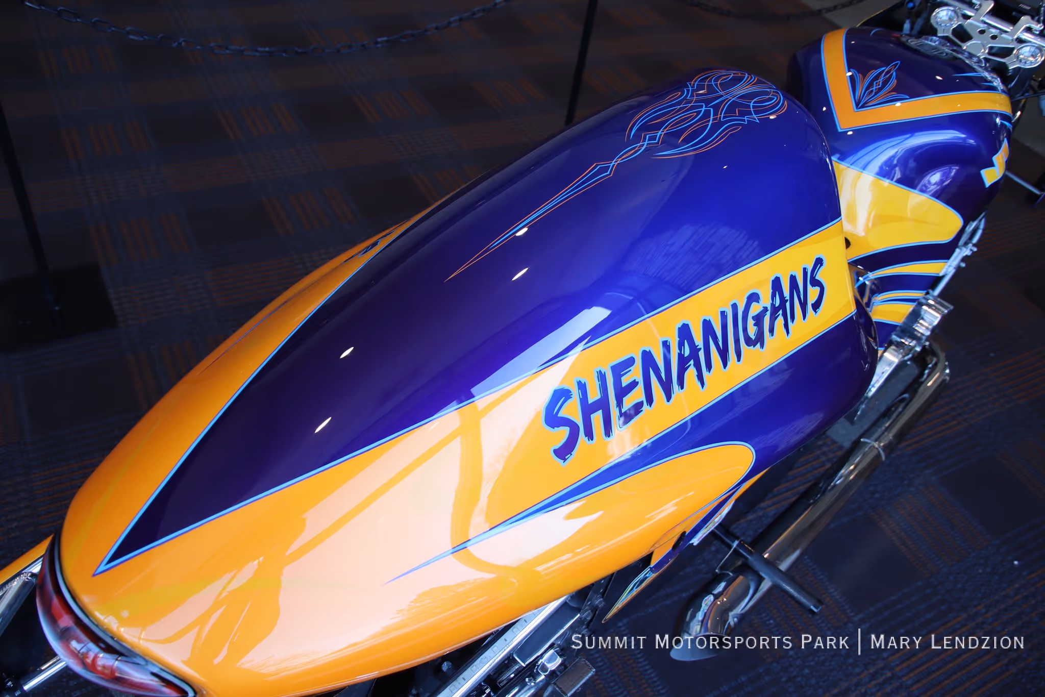 Close-up of a custom motorcycle gas tank with purple and yellow paint and the word 'SHENANIGANS' in blue letters.