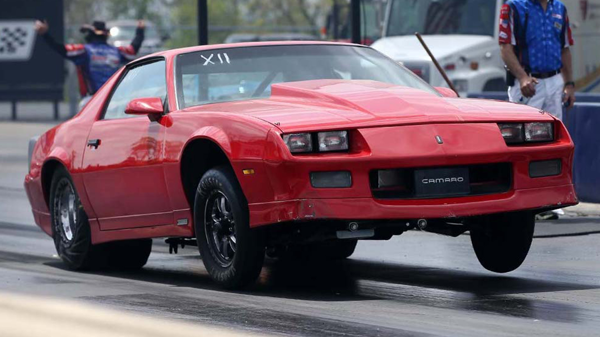 Red Chevrolet Camaro with front wheels lifted off the ground during a drag race.
