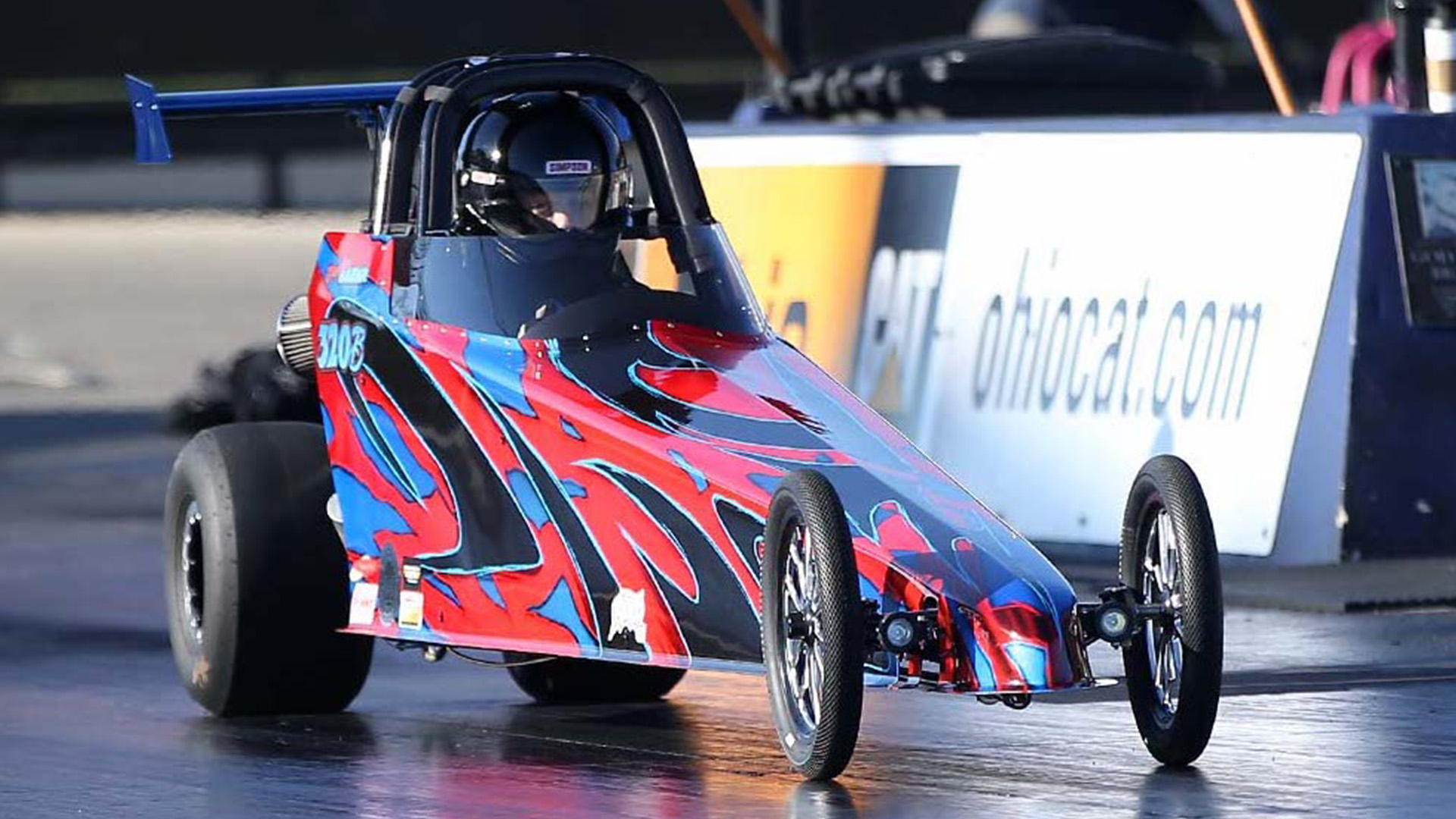 Colorful drag racing car with red, blue, and black design on a race track, driver visible wearing a helmet.