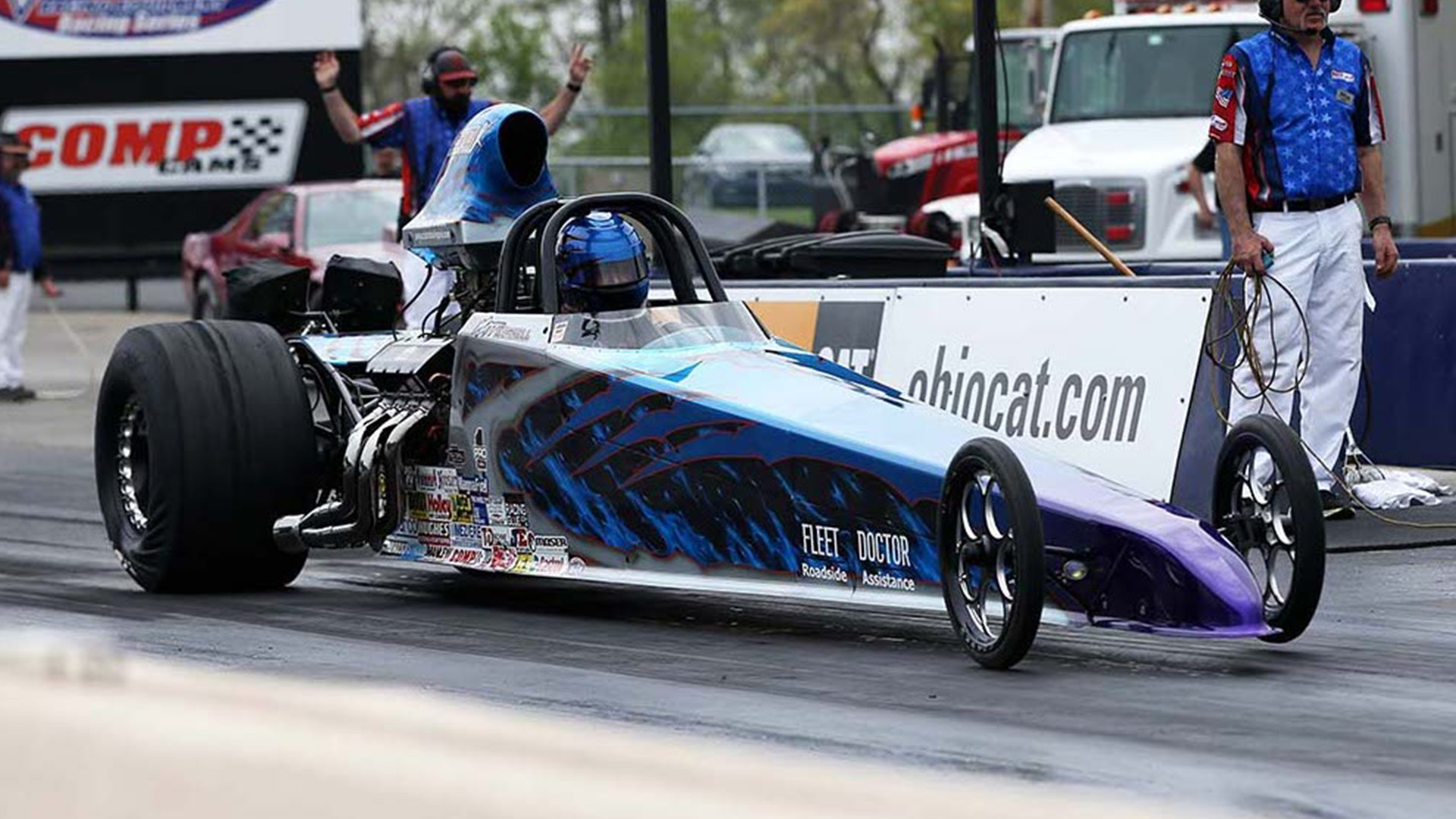 Blue and purple dragster car on a racetrack with driver in helmet and crew members in background.