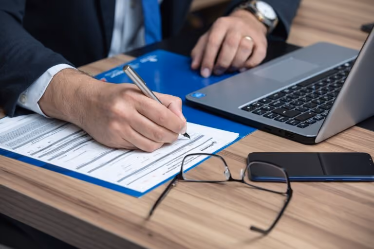 Person in business attire signing a document on a blue clipboard beside a laptop, with glasses and a smartphone on the wooden desk.