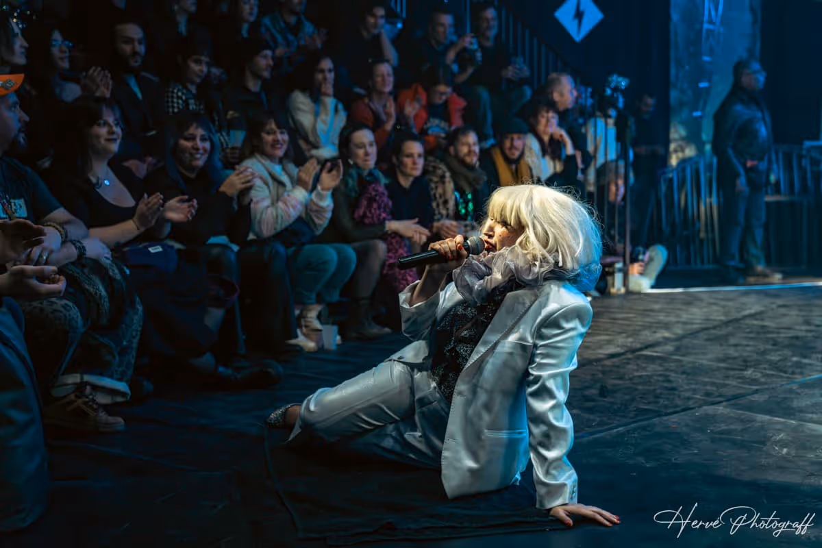 Chanteuse en costume clair assise au sol avec un micro, face au public qui applaudit.