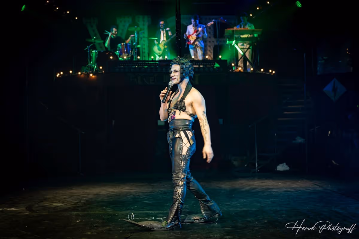 Performeur en pantalon de cuir décoré, tenant un micro sur scène devant les musiciens.