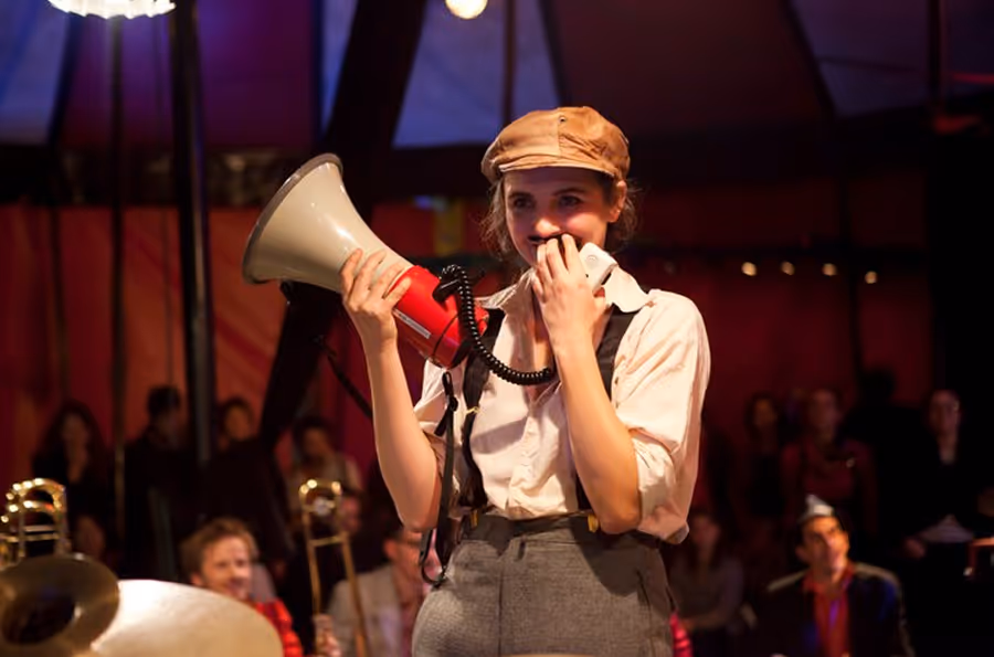 Cirque Électrique Paris spectacle - artiste femme avec mégaphone rouge et blanc, casquette beige, bretelles, pantalon gris, ambiance chapiteau, public en arrière-plan, performance théâtrale, scène intimiste, maîtresse de cérémonie, arts du cirque contempor