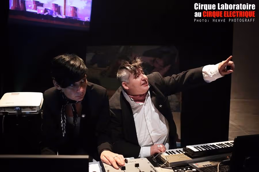 Cirque Électrique Laboratoire Paris performance musicale - duo artistes avec instruments électroniques, table de mixage, costume élégant noir et blanc, spectacle audiovisuel expérimental, projection vidéo, musique électronique alternative.